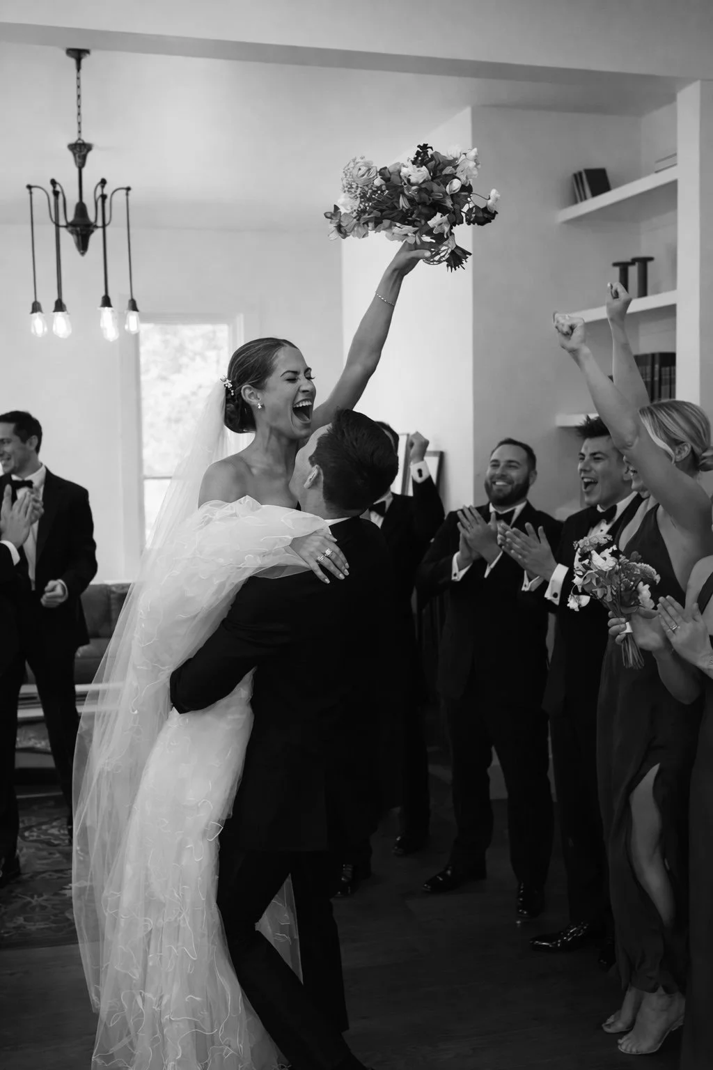 A newlywed couple in wedding attire celebrating outside a cream-colored building, with the groom opening a champagne bottle, and the bride smiling and holding his arm, surrounded by green bushes.
