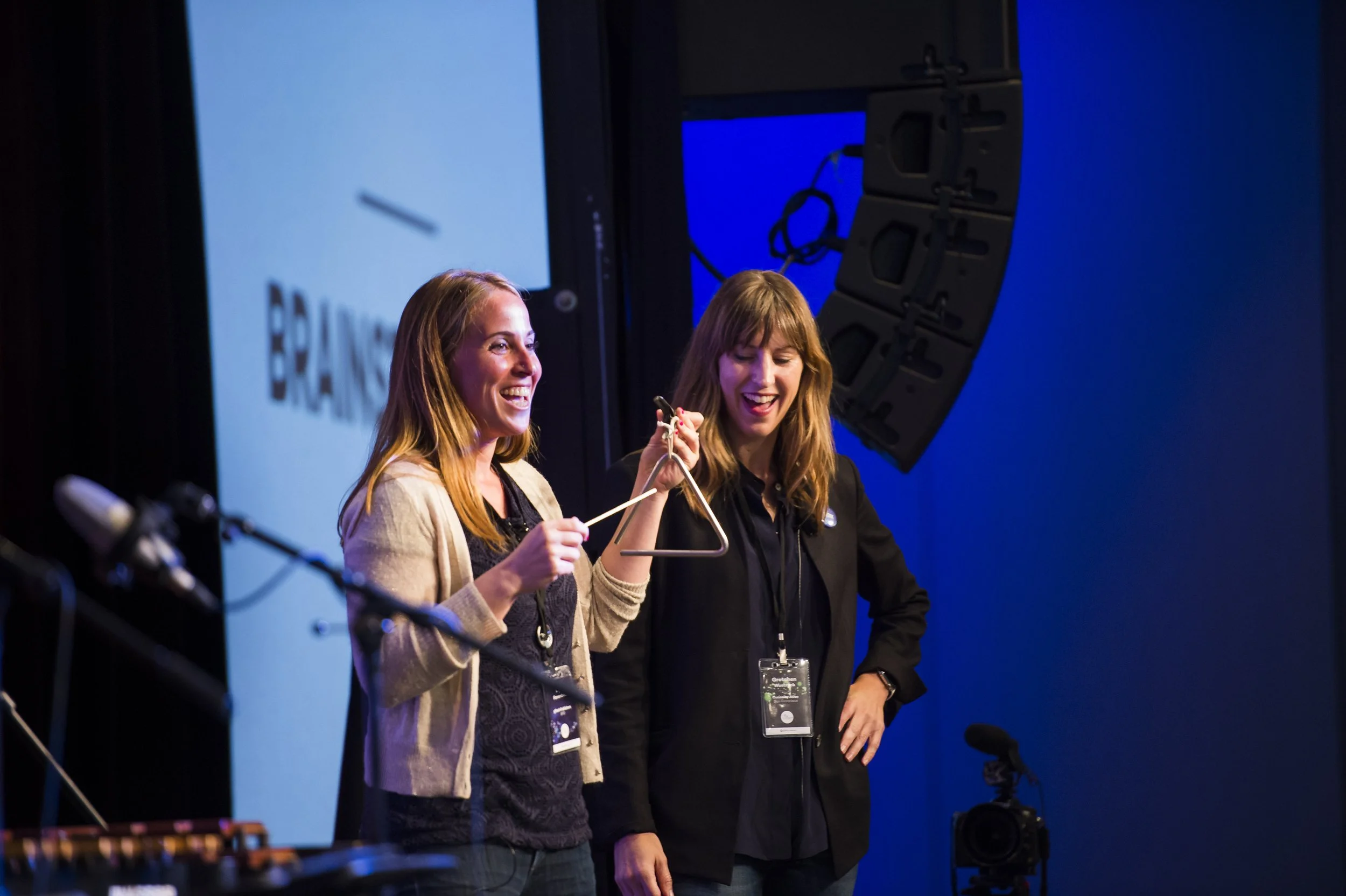 Two women smiling and talking on stage at a conference or event, with large screens and audio equipment in the background.