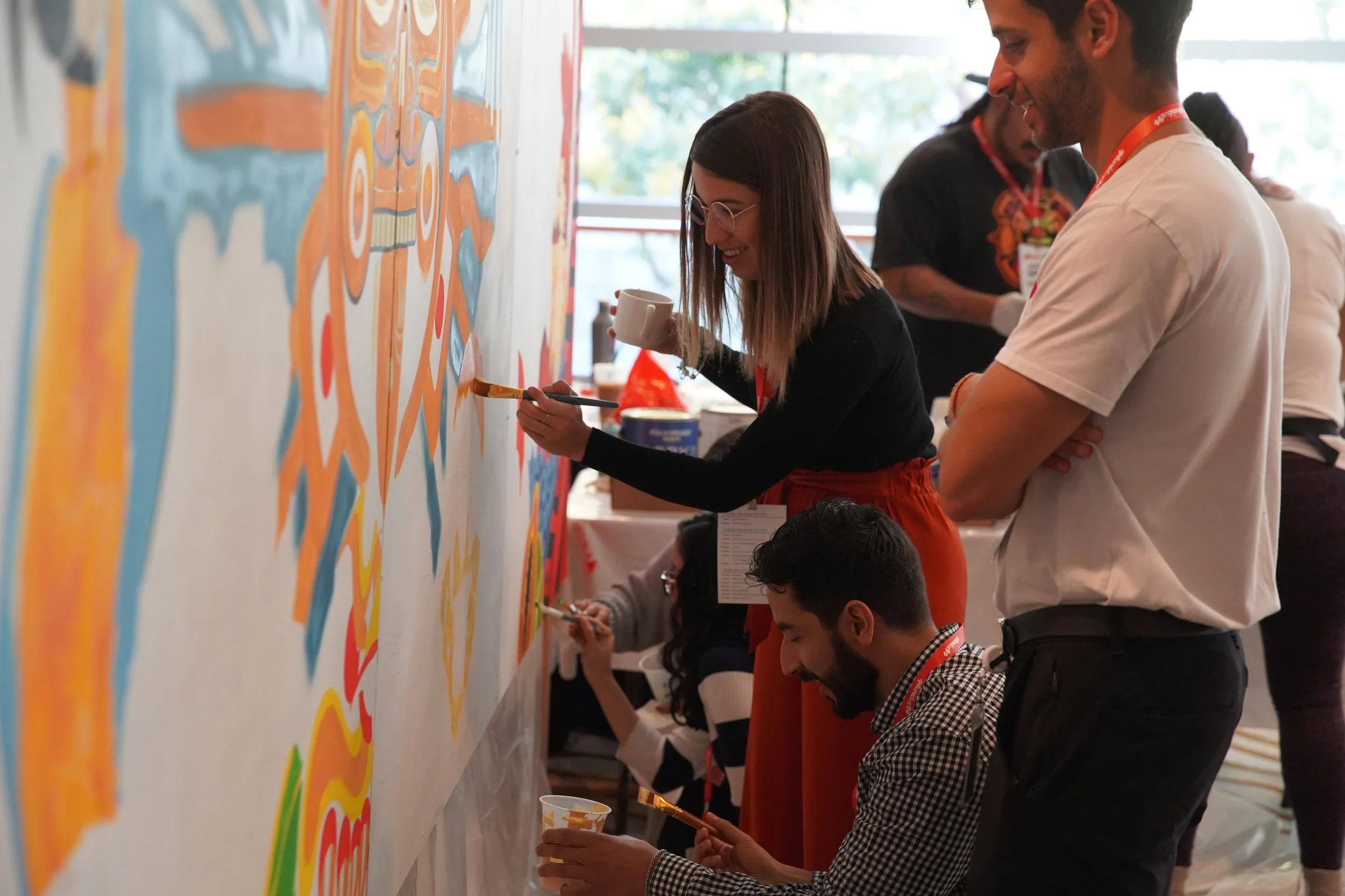 People painting a colorful mural on a wall at an indoor event.