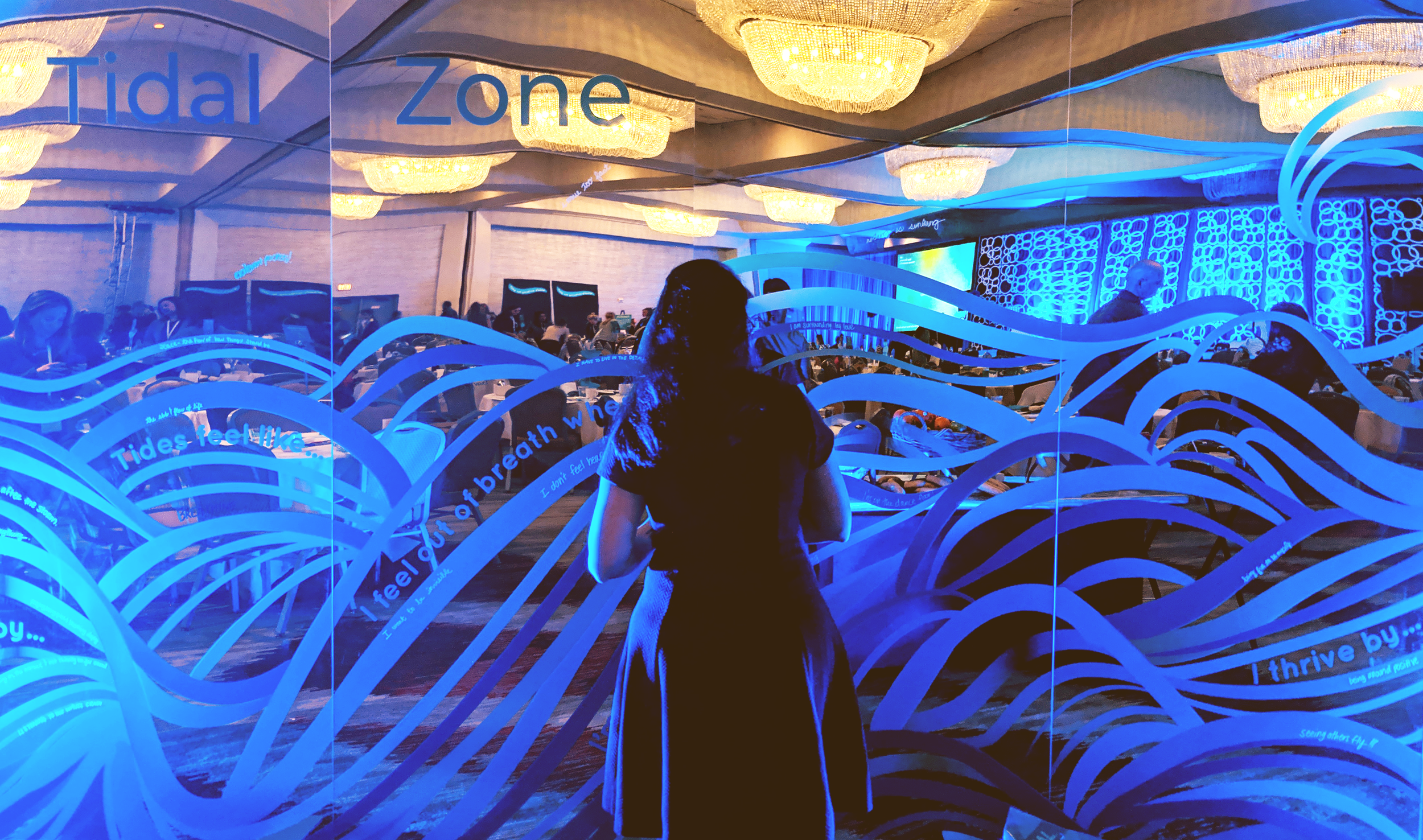 A woman standing in front of a glass wall decorated with blue wave patterns and writing. Behind her, there is a large event space with chandeliers, people sitting at tables, and a screen on the wall. The room is dimly lit with blue lighting.