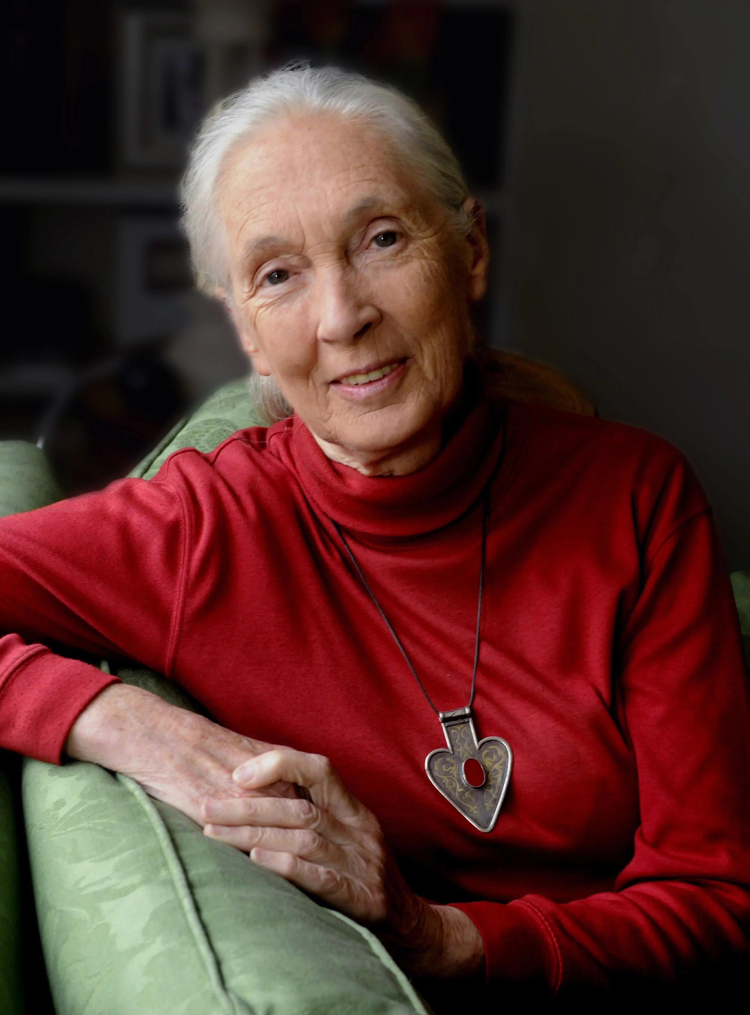 Jane Goodall wearing a red long-sleeve shirt and a large key-shaped necklace, sitting in a chair, smiling softly, with a blurred background.