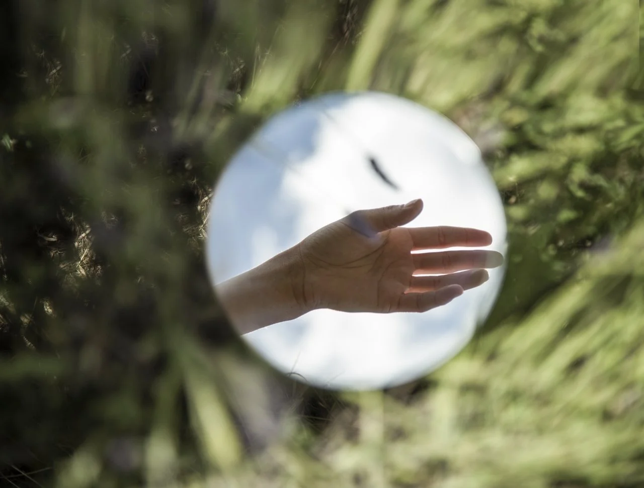 A hand extended through a circular mirror reflecting the sky and clouds, with green foliage around the mirror.