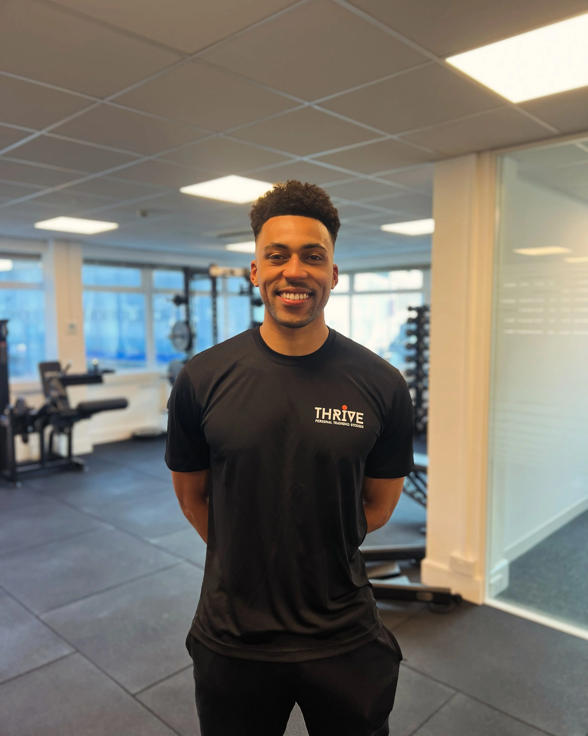 A smiling young man with curly hair, wearing a black Thrive personal training store t-shirt, standing in a gym with exercise equipment in the background.