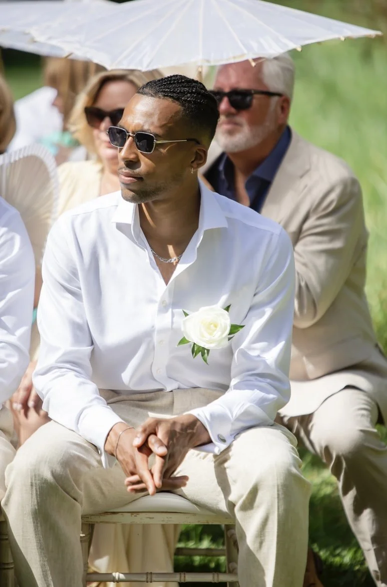 A group of people attending an outdoor wedding ceremony during daytime. The man in the foreground is wearing sunglasses, a white shirt with a white flower boutonniere, and beige pants. He is sitting with his hands clasped and appears to be focused. Behind him, there are other guests, some with sunglasses, sitting under a translucent white umbrella. The background features green grass and blurred natural scenery.