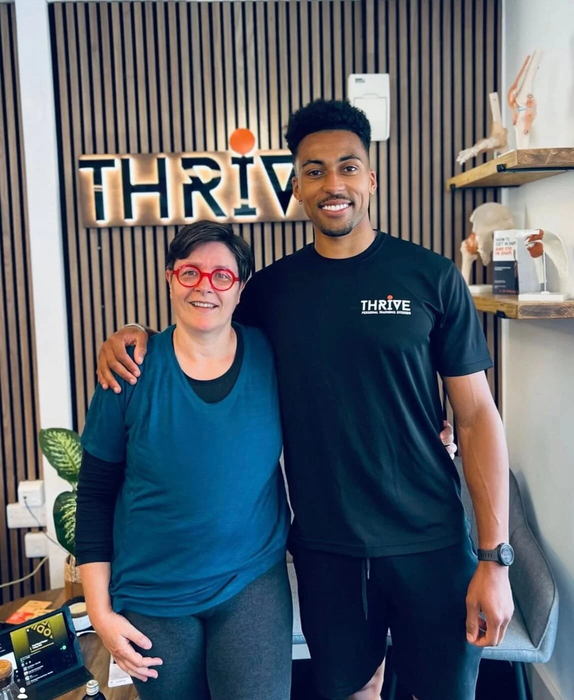 A woman and a man standing together inside a room, smiling for the camera. The woman has short dark hair, red glasses, and is wearing a blue shirt. The man has short curly hair, a watch, and is wearing a black shirt with the word 'THRIVE' on it. Behind them is a wall with a sign that says 'THRIVE' and some shelves with decorative items.