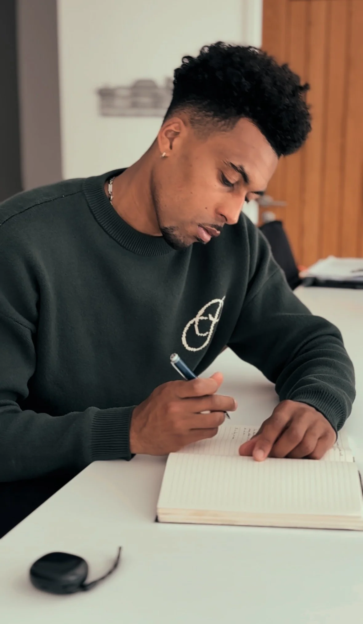 A young man sitting at a desk, writing in a notebook with a pen, while wearing a dark green sweatshirt with a white logo on it.