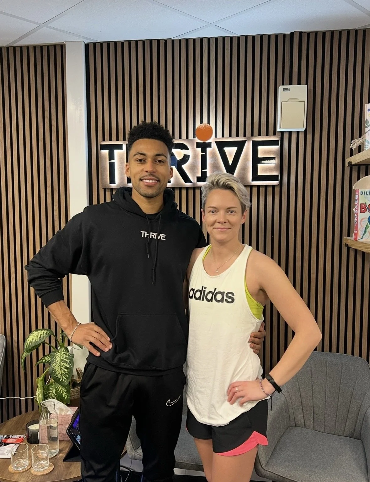 A man and woman stand together in a room with a wooden slat wall and a sign that reads 'THRIVE' behind them. The man is wearing a black hoodie and pants, and the woman is in a white tank top and black shorts with pink trim. The room has a small table with water bottles, glasses, and a plant.
