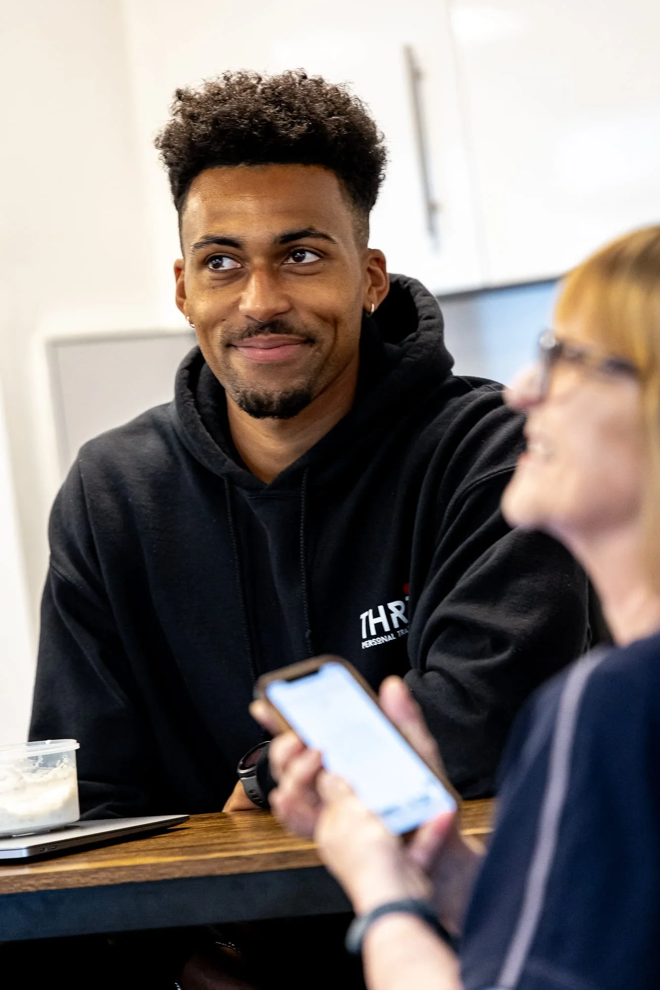 A young man with curly hair, earrings, and a goatee smiling while sitting at a table with a woman holding a smartphone. The woman has blond hair, glasses, and is smiling as they converse.
