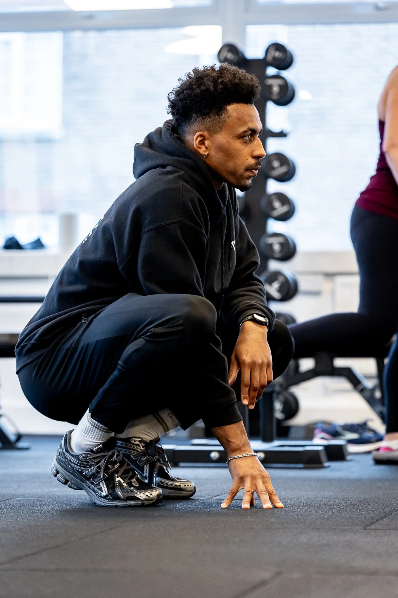 A man crouching on the gym floor in workout attire, with dumbbells in the background.