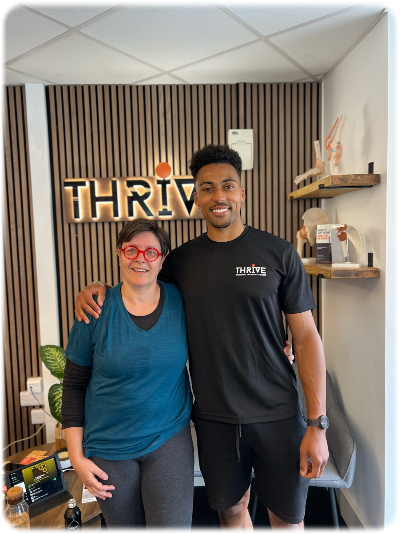 A woman with glasses and a man in a Thrive shirt standing together indoors, smiling, in front of a wooden wall with the Thrive logo and shelves with decor items.