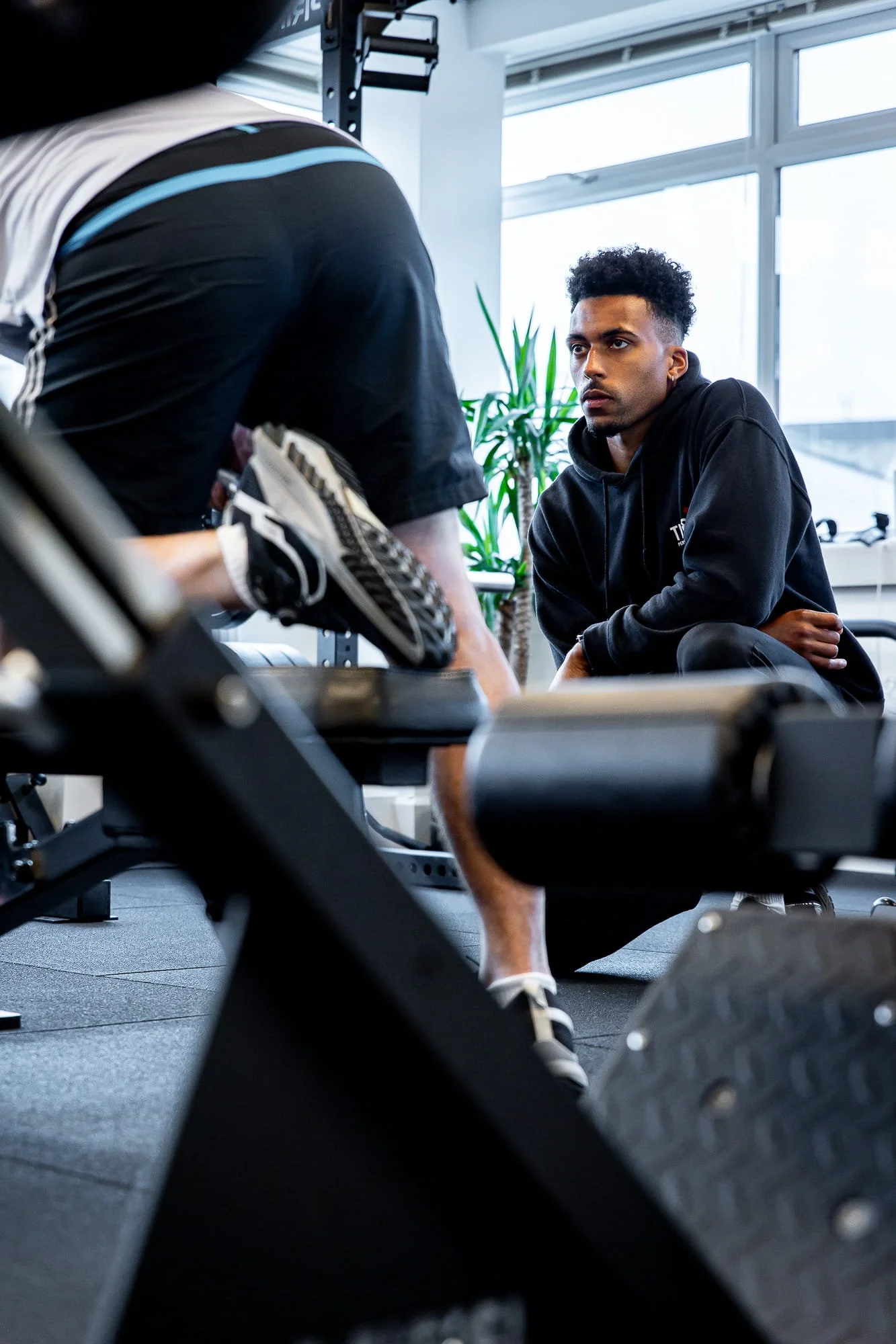 A young man in a black hoodie sitting and listening to a person in gym clothes talking during a workout session at a gym.