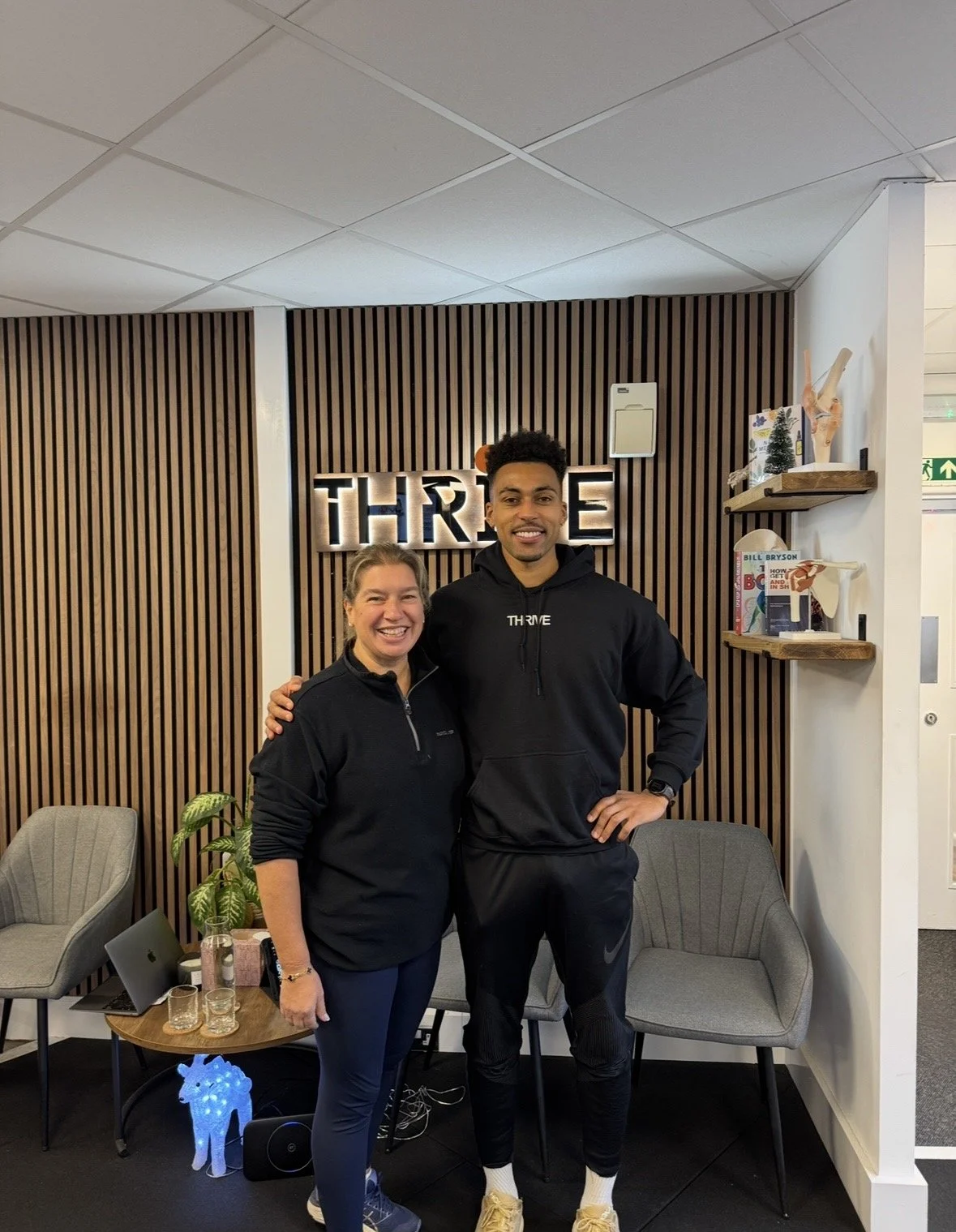 Two smiling people, a woman and a man, standing together indoors in front of a wooden slat wall with illuminated "THRIVE" sign, surrounded by chairs, a small table, shelves with books and decorations.
