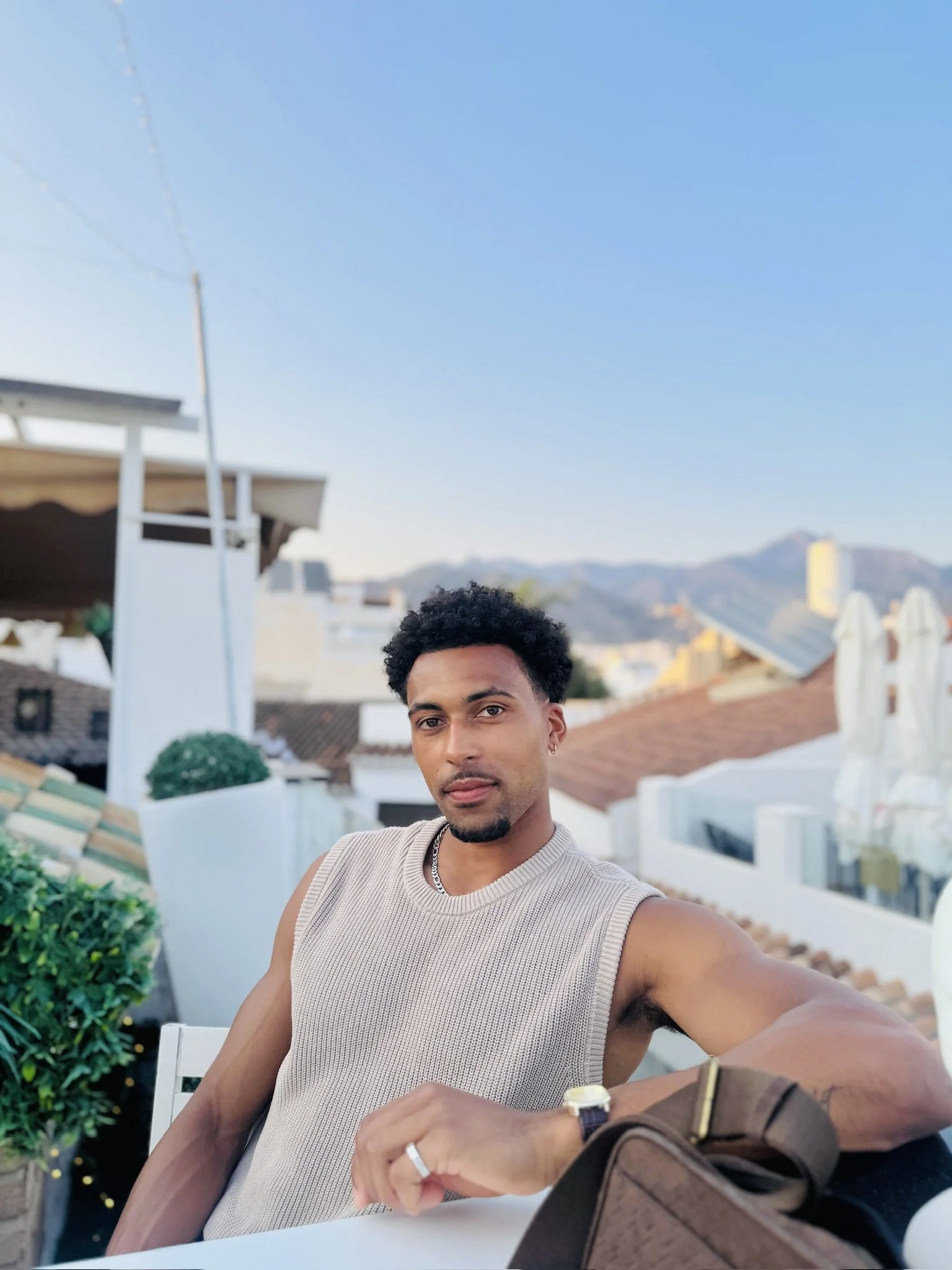 A young man with curly hair, wearing a sleeveless beige shirt and jewelry, taking a selfie outdoors at a rooftop or terrace. The background includes a clear blue sky, rooftops, and a mountain in the distance.