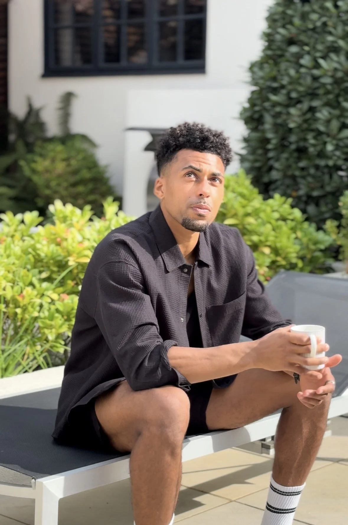 A young man with curly black hair and a goatee sitting on a lounge chair outdoors, holding a white mug, wearing a black shirt, shorts, and striped socks, surrounded by greenery and a house in the background.