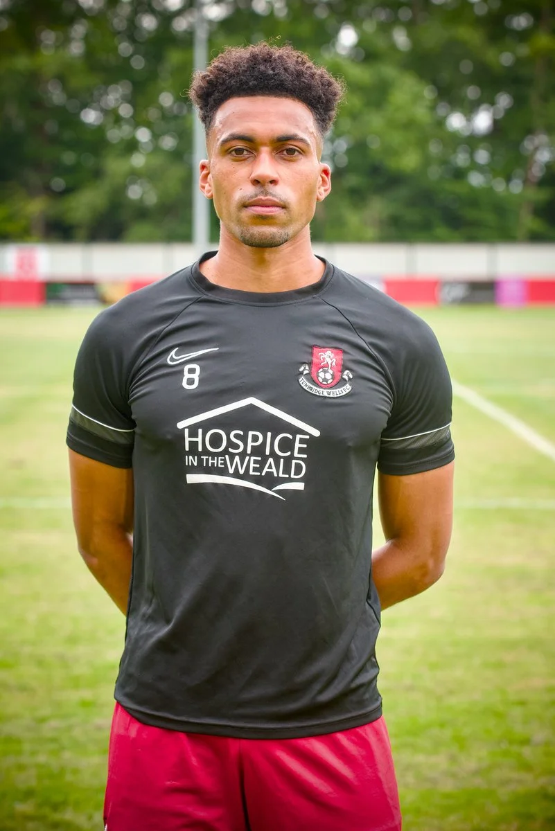 A young man standing on a soccer field wearing a black jersey with the number 8, red shorts, and standing with his hands behind his back, with trees and a fence in the background.
