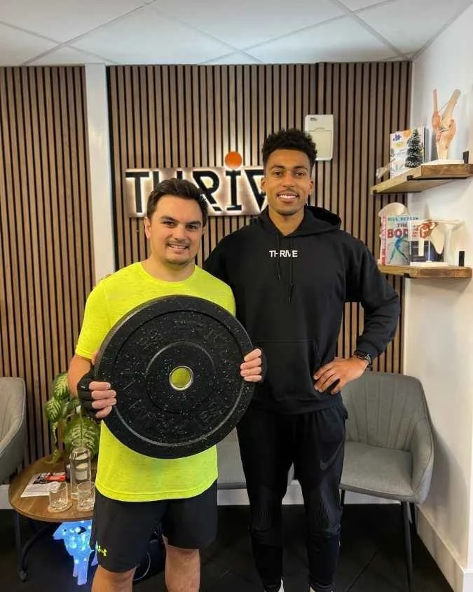 Two men standing inside a fitness or gym facility, one holding a black weight plate, and the other with his hand on his hip, smiling. Behind them is a wall with a logo that says 'TRIVE'. There are shelves with books and decorative items on the right and a small table with glasses and a plant on the left.