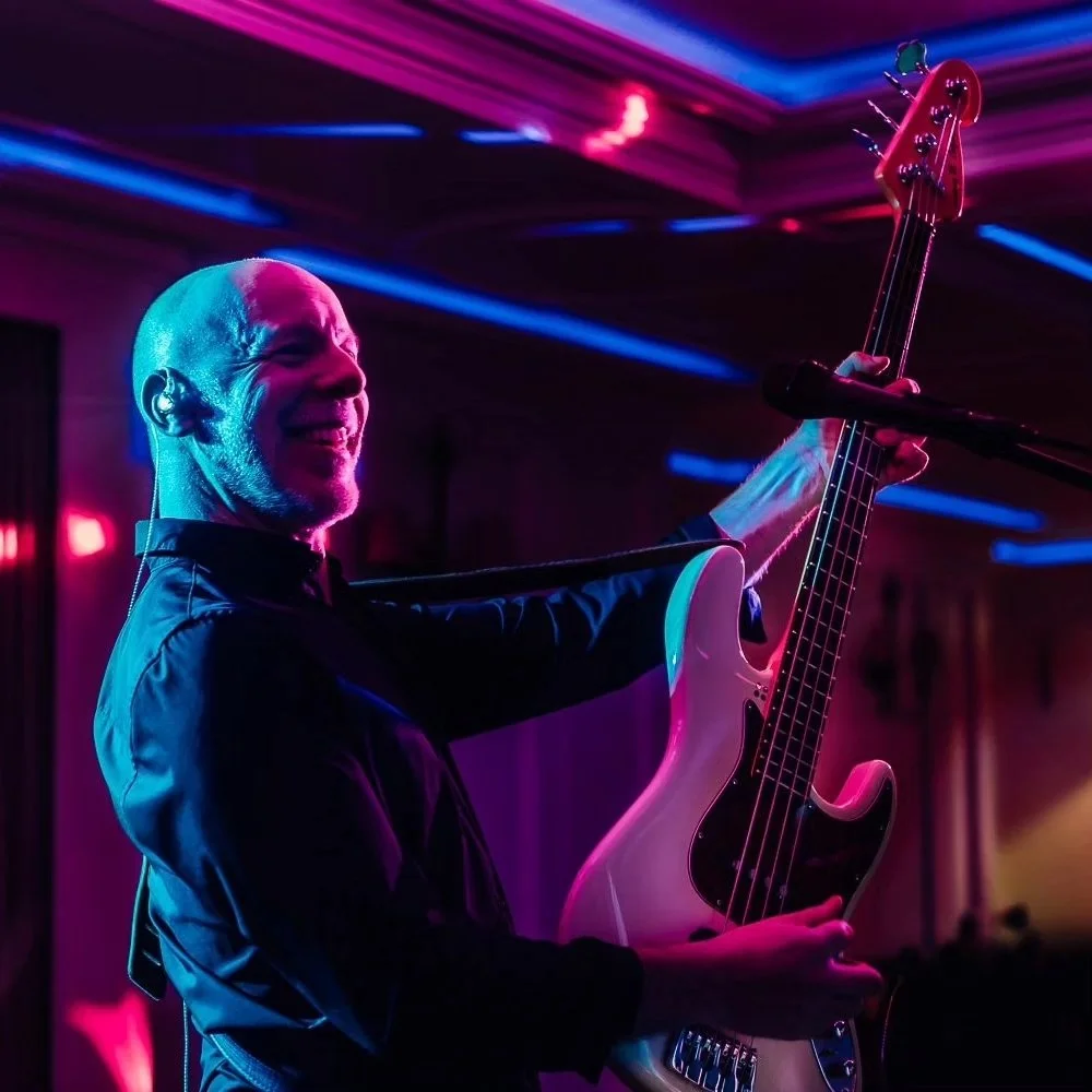 A man with a shaved head playing an electric bass guitar in a colorful, neon-lit setting, smiling and enjoying the music.