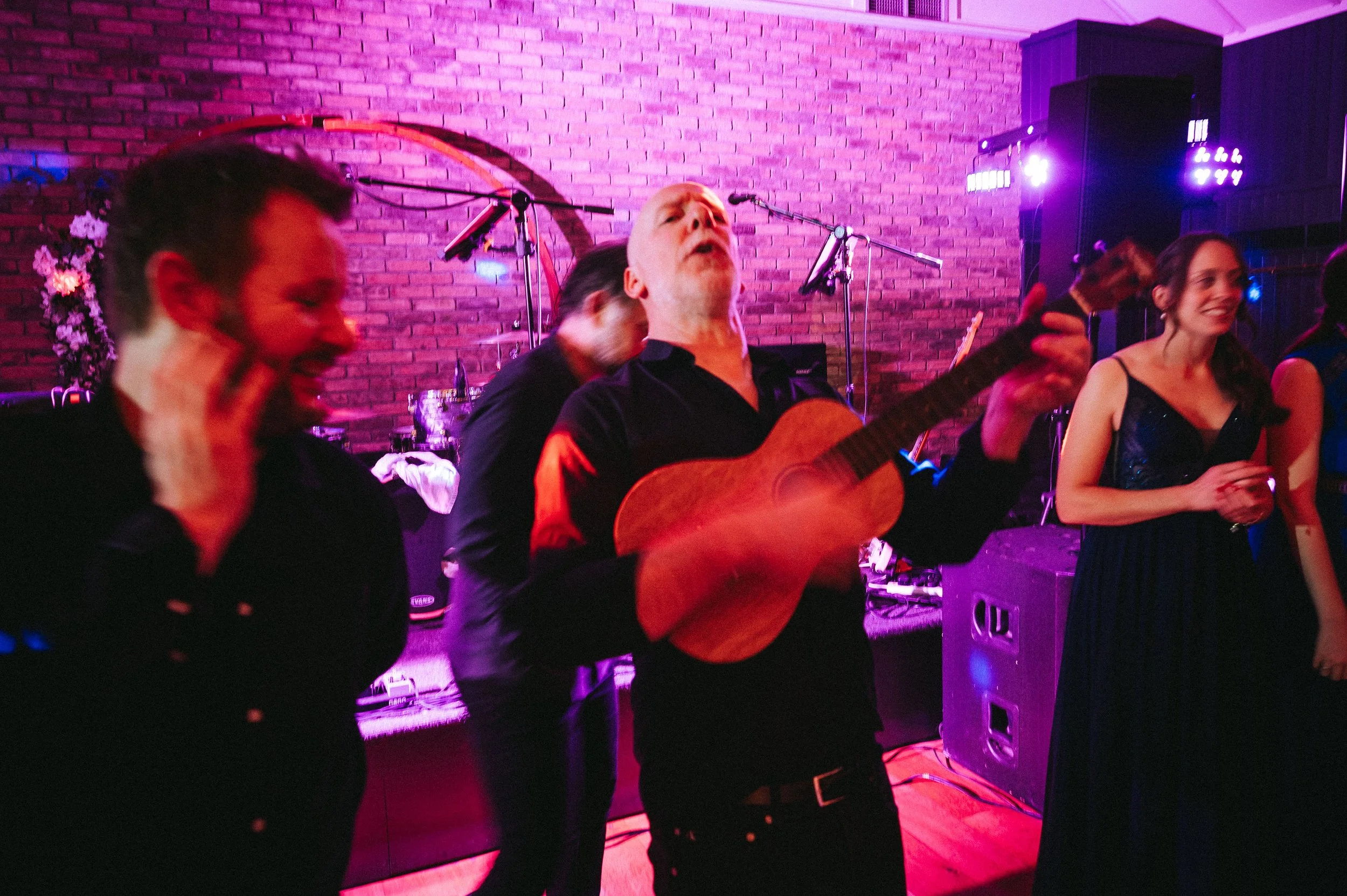 People enjoying live music at a venue with brick walls and colorful stage lighting. One man is playing guitar with his eyes closed, others are smiling and dancing.