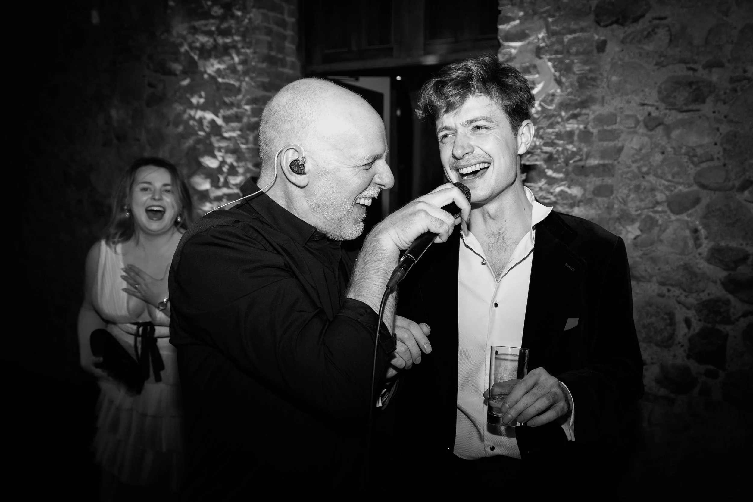 Two men singing into a microphone at a lively party or event, with a woman laughing in the background. Black and white photo.