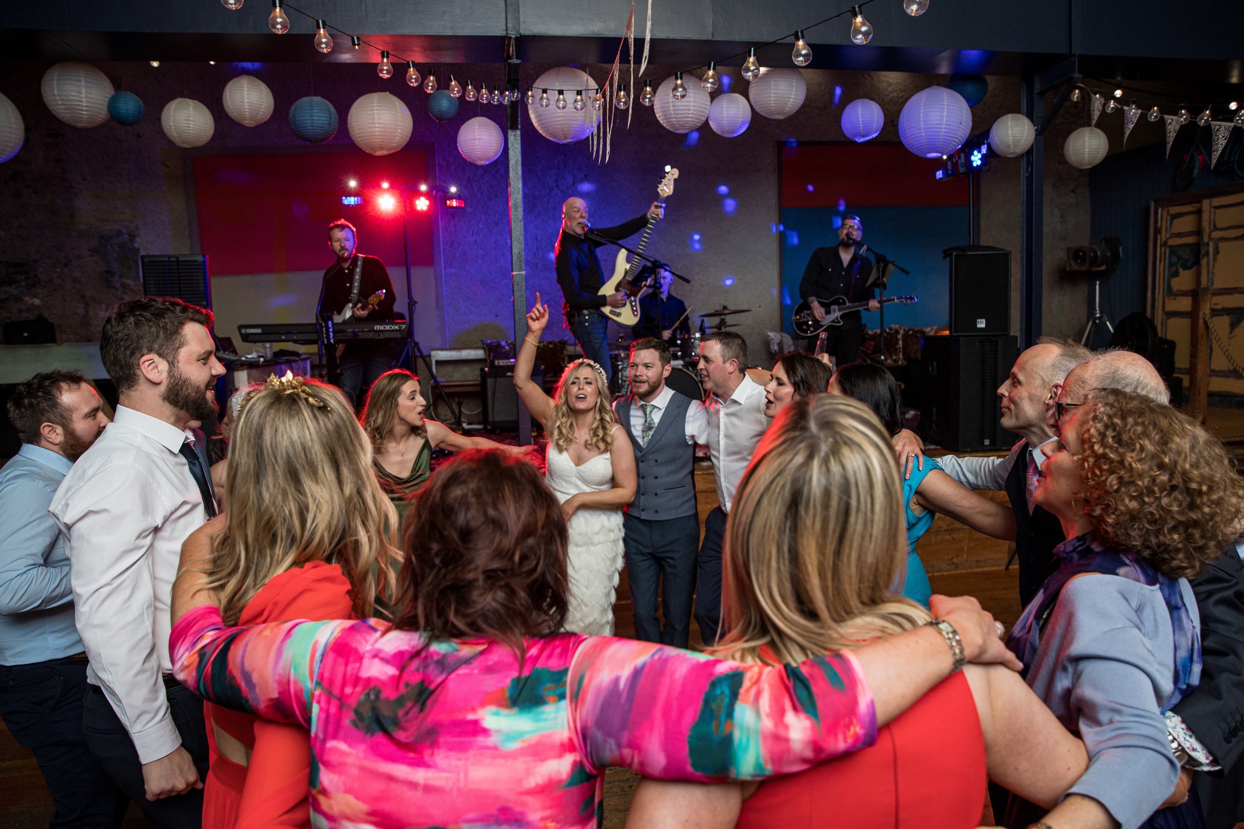 People dancing and celebrating at a wedding reception with a live band performing on stage in the background. The bride and groom are at the center, surrounded by friends and family, all enjoying the lively atmosphere.