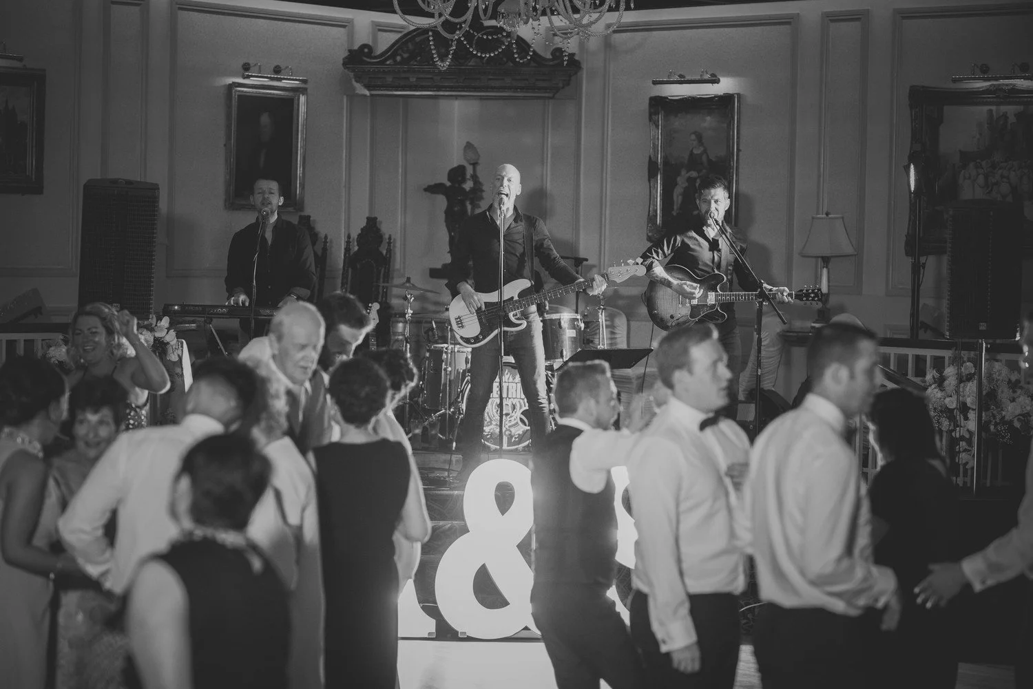 A black-and-white photo of a band performing on stage at a formal event. The band includes a drummer, a bassist, and two guitarists, with guests dancing in front of the stage. The event takes place in a decorated room with portraits on the wall and a chandelier hanging from the ceiling.