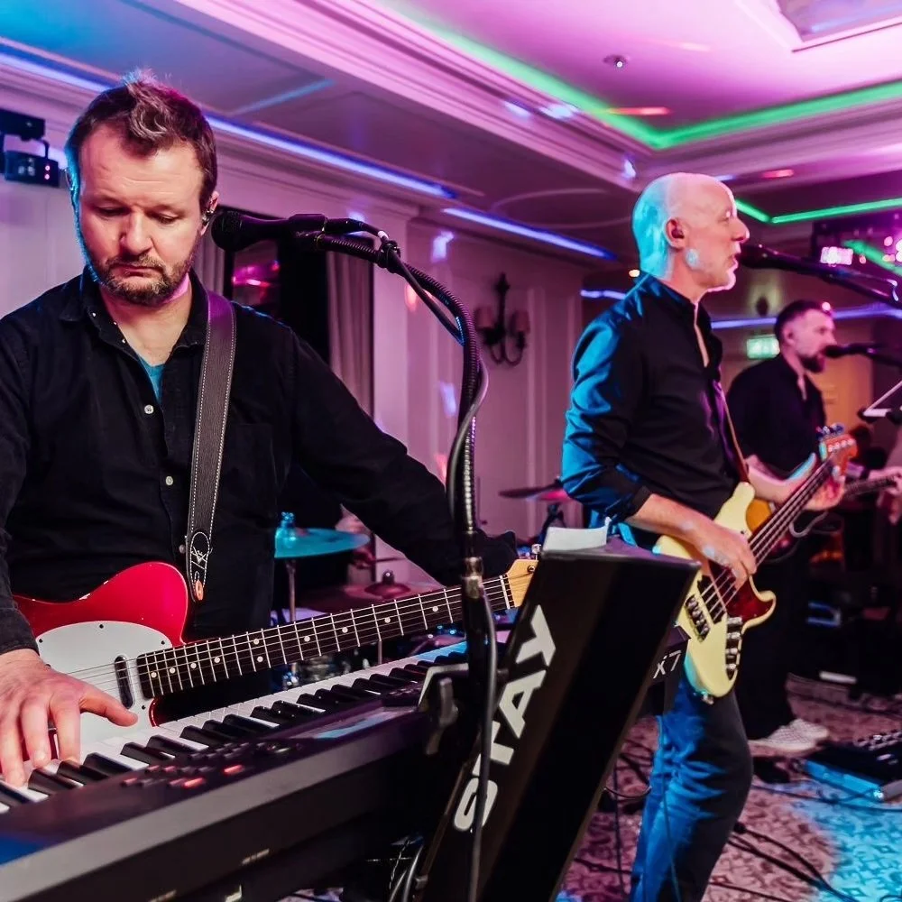Three musicians performing at an indoor event with colorful stage lighting, including a keyboard player, a bassist, and another guitarist.