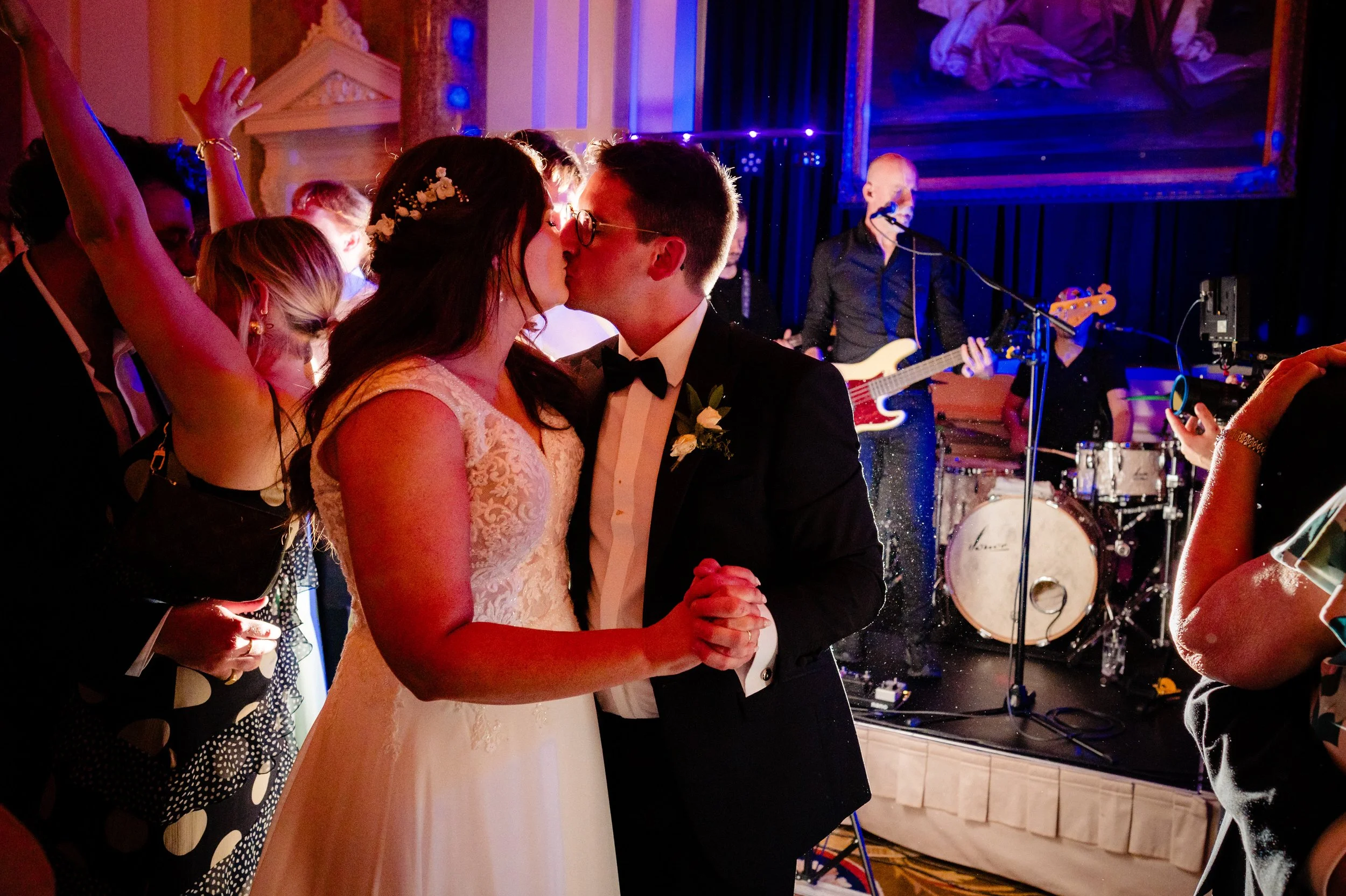 A bride and groom share a kiss during their wedding reception. The bride wears a lace wedding dress with floral hair accessories, and the groom is in a black tuxedo with a bow tie. Guests surround them, some raising their hands. A live band plays in the background with musicians and instruments visible, including a guitarist and drums.