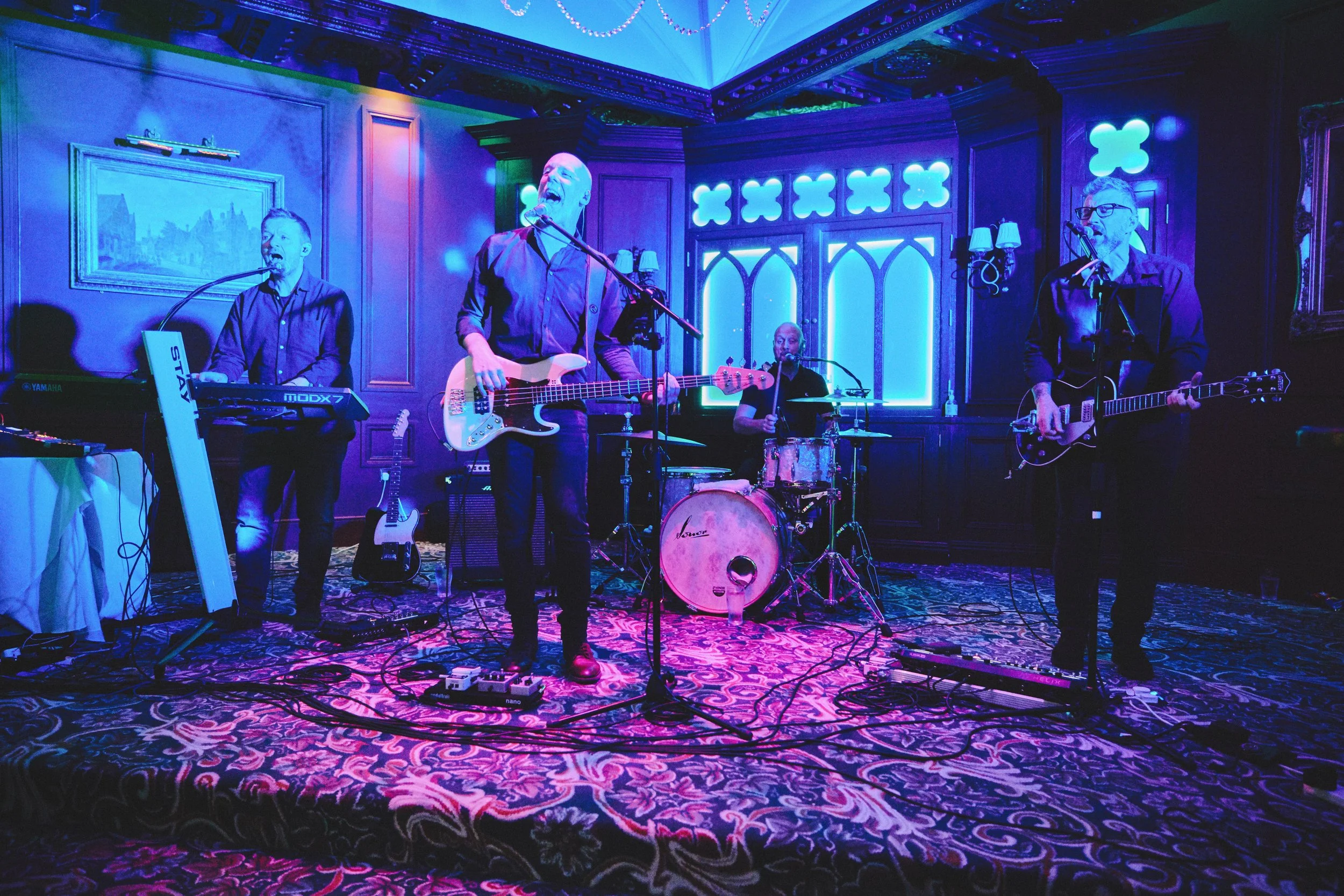 A band performing live on a stage with purple and blue lighting in a cozy, decorated room with wooden paneling, a painting, and illuminated decorative shapes on the wall. The band consists of five members: a keyboardist, a bassist, a drummer, and two guitarists, all singing into microphones.