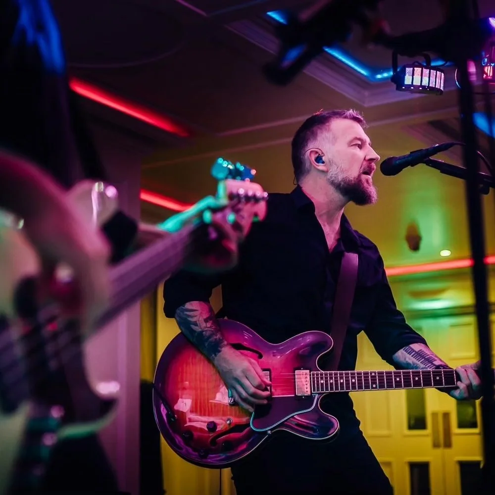 A musician playing an electric guitar and singing into a microphone at a live performance. The background has colorful lighting in a dimly lit room.