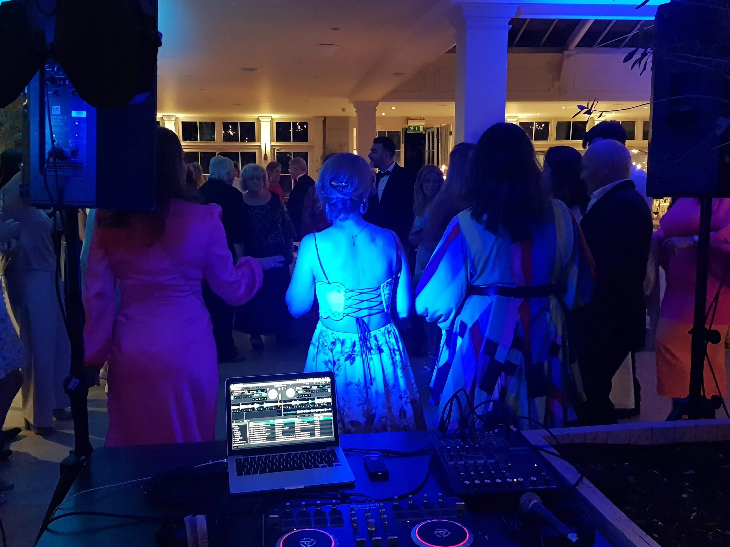 People dancing and socializing at a party or wedding reception with DJ equipment and lighting.