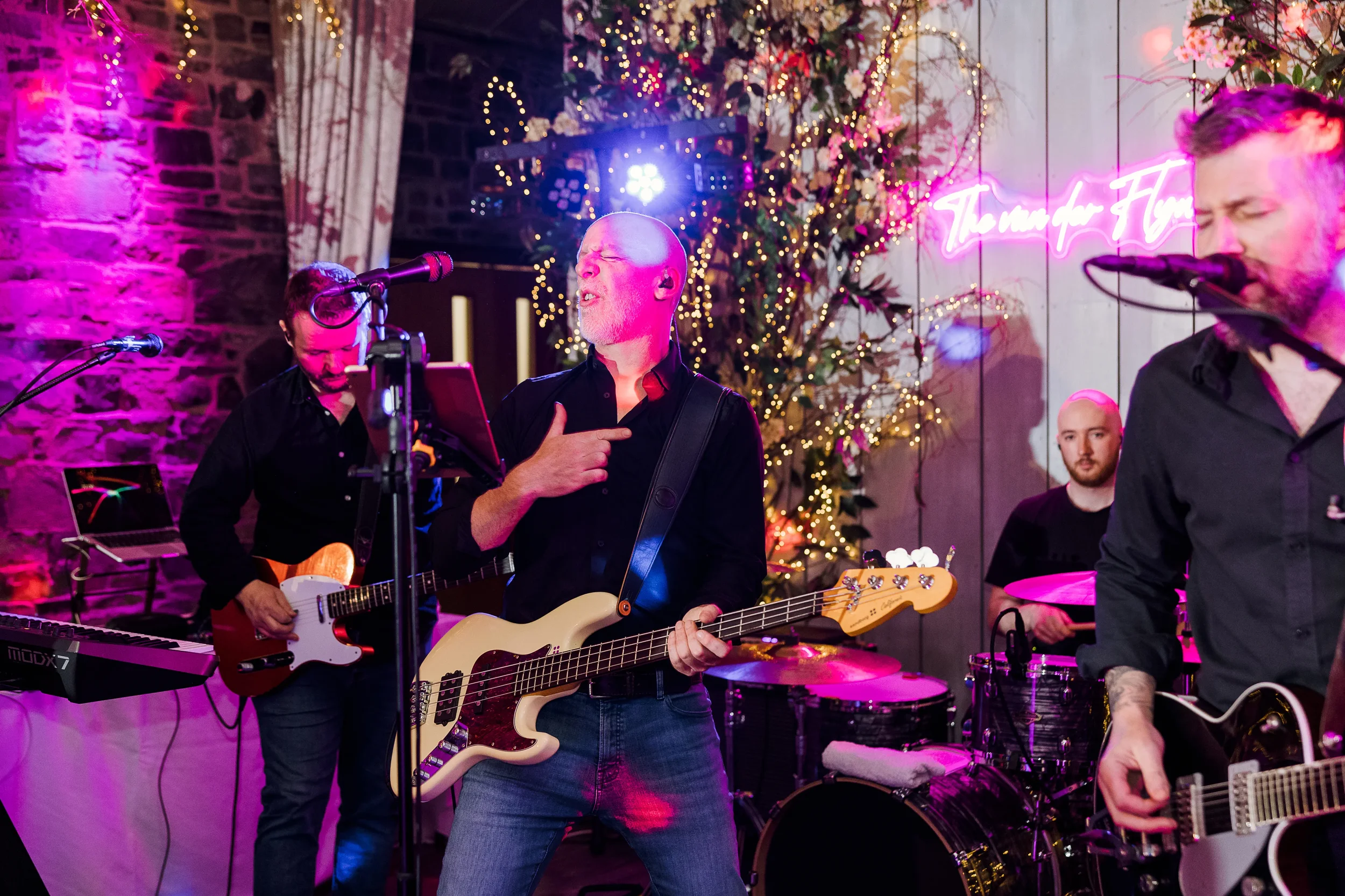 A band of four musicians performing at a decorated indoor venue with Christmas lights and a neon sign that reads "The meaning of Humor." The lead singer in the center plays a bass guitar, singing with eyes closed and hand on chest. To his left, a guitarist with a small red electric guitar smiles. On the far right, another guitarist and vocalist plays an electric guitar. In the background, a drummer plays drums.