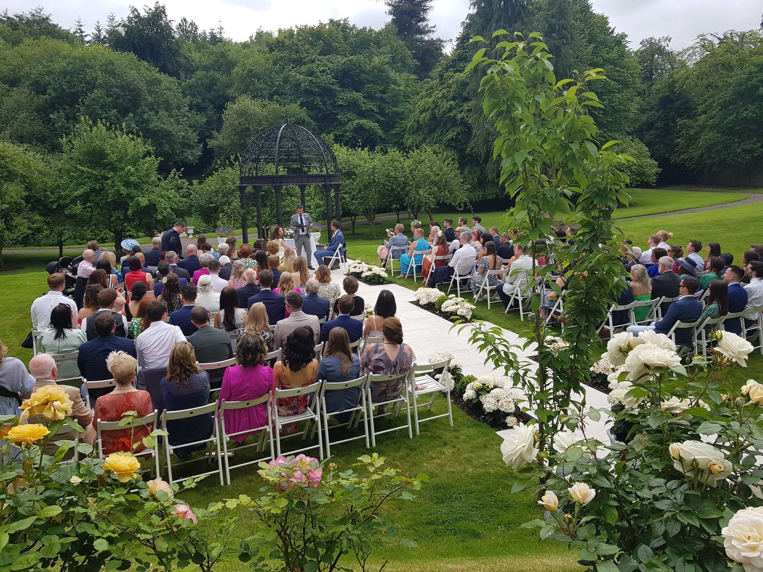 Outdoor wedding ceremony with guests seated under a gazebo in a lush green garden, surrounded by flowering bushes.