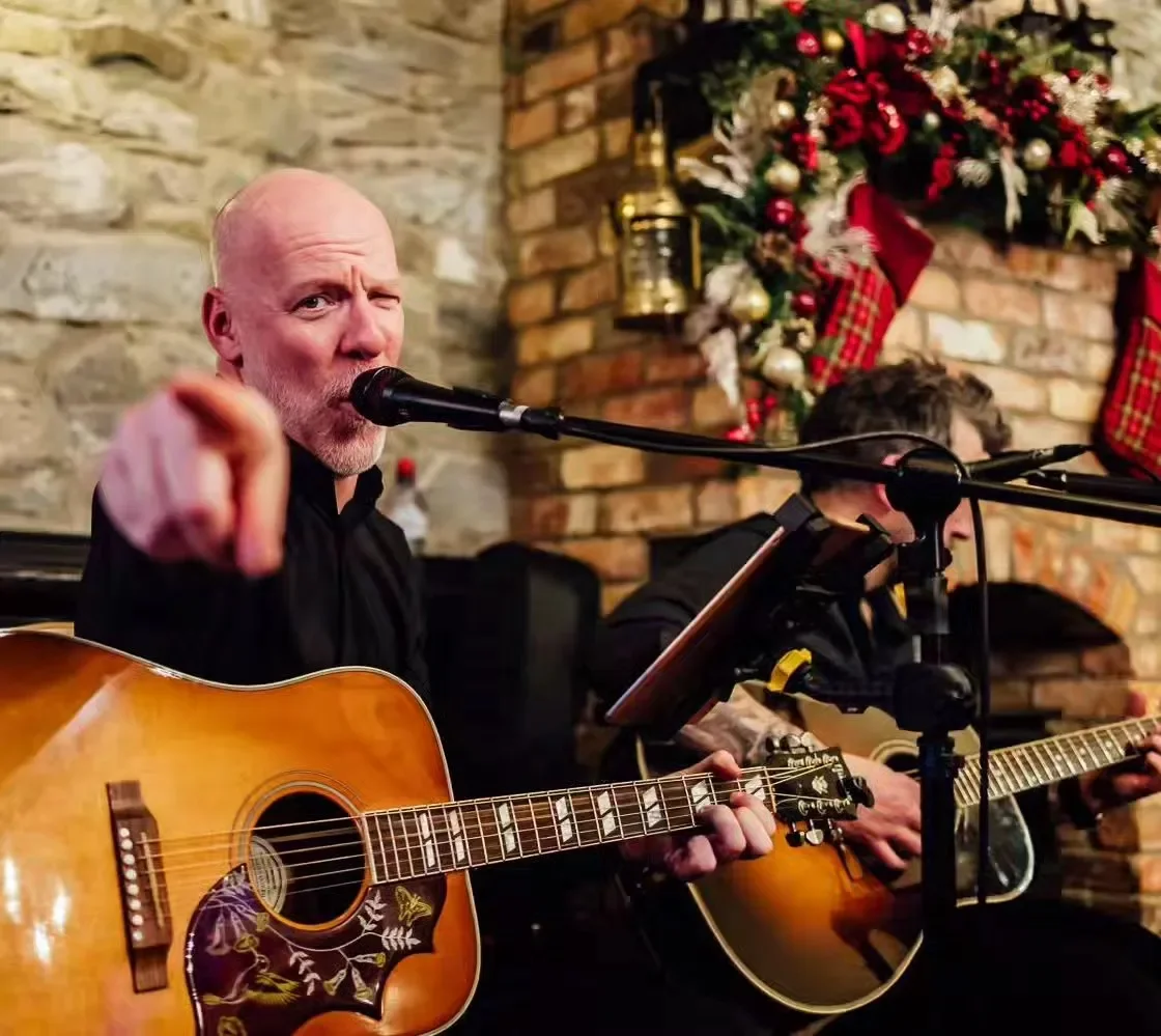 A man with a bald head and beard pointing at the camera while singing into a microphone and playing an acoustic guitar, with Christmas decorations including stockings and a wreath in the background.