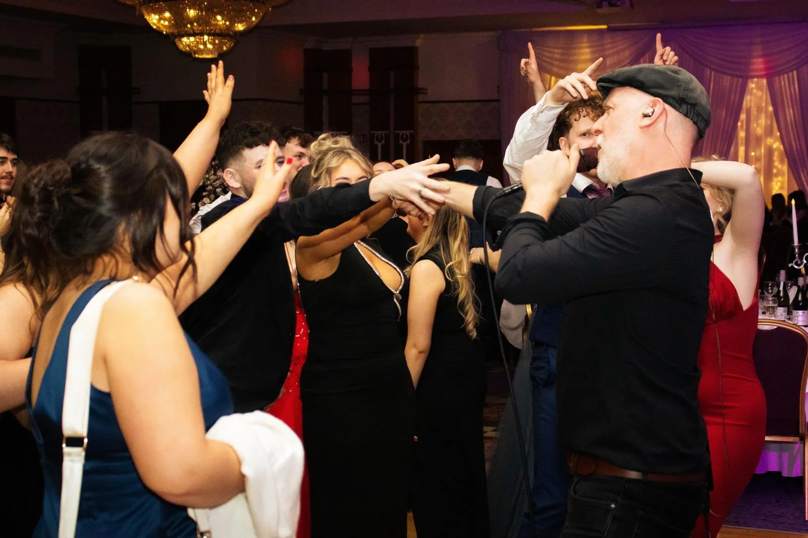 A singer with a gray beard and wearing a black shirt and cap performs at a lively party or celebration, surrounded by people dancing and singing along in an elegant venue with chandeliers and purple drapes.