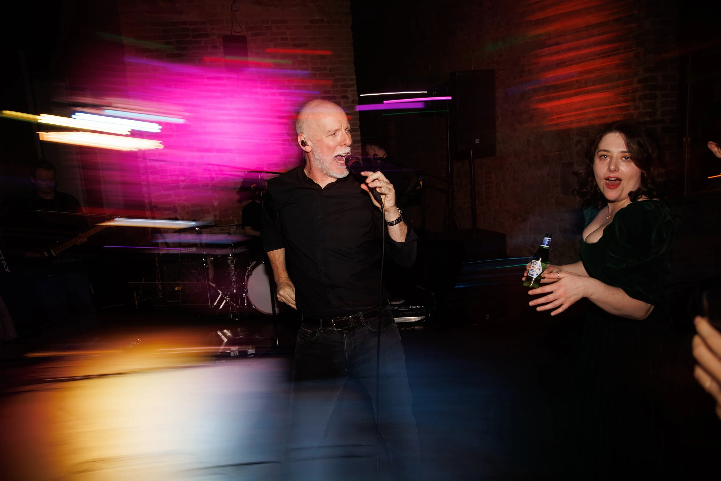A lively indoor party scene with a man singing into a microphone and a woman holding a beer bottle, with colorful motion effects and a brick wall background.