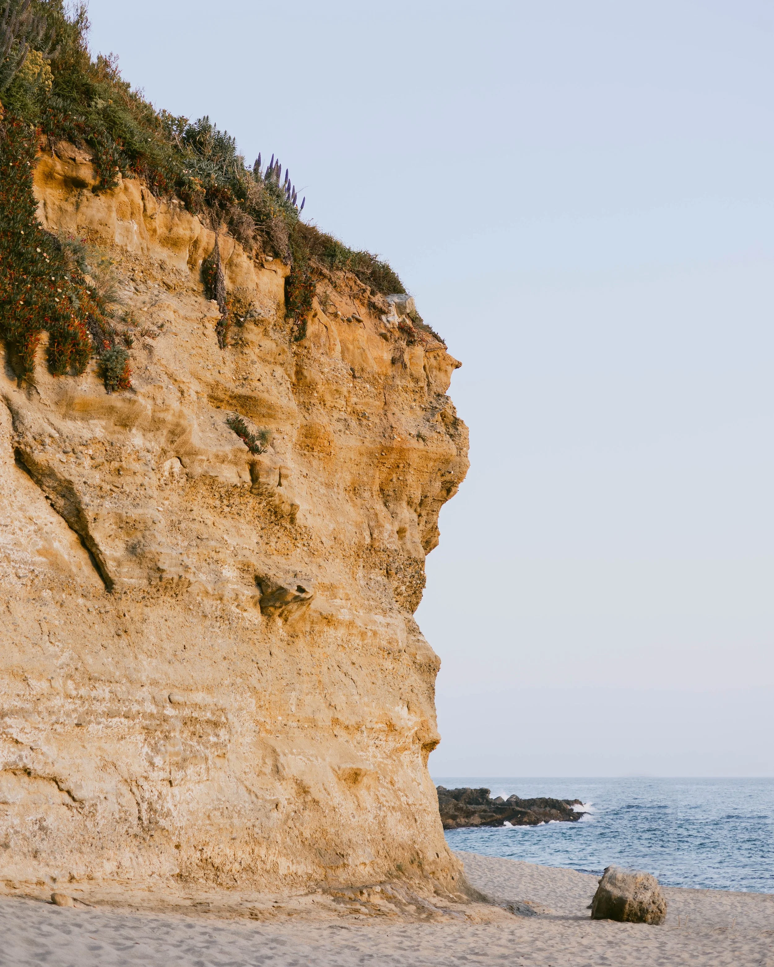 Cliffside at the beach with sand, rocks, and ocean water visible below, under a clear sky.