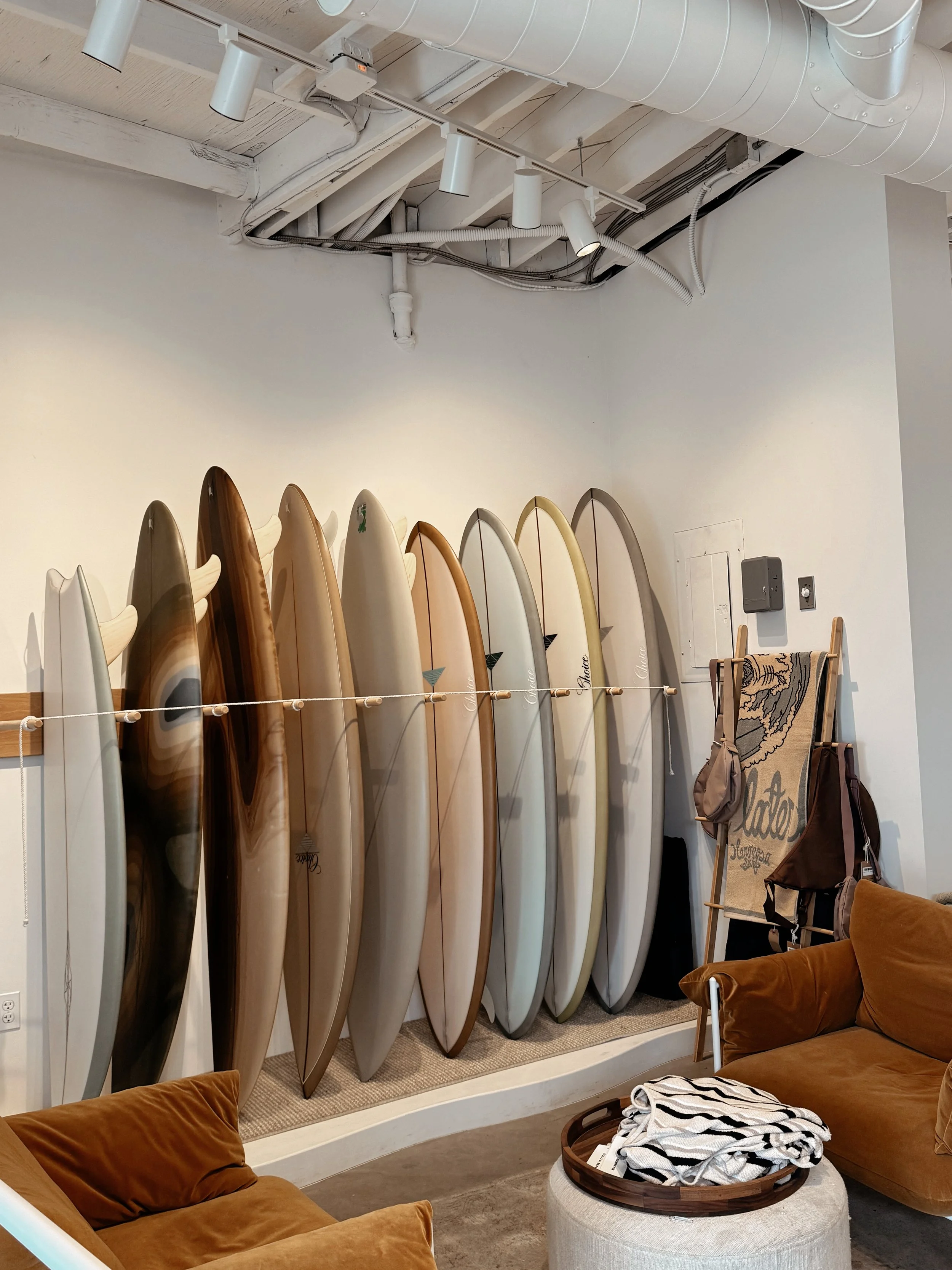 Interior of a surf shop with multiple surfboards standing upright against a wall, a brown sofa, a folding chair with a towel draped over it, and a round ottoman with a striped cloth on top.
