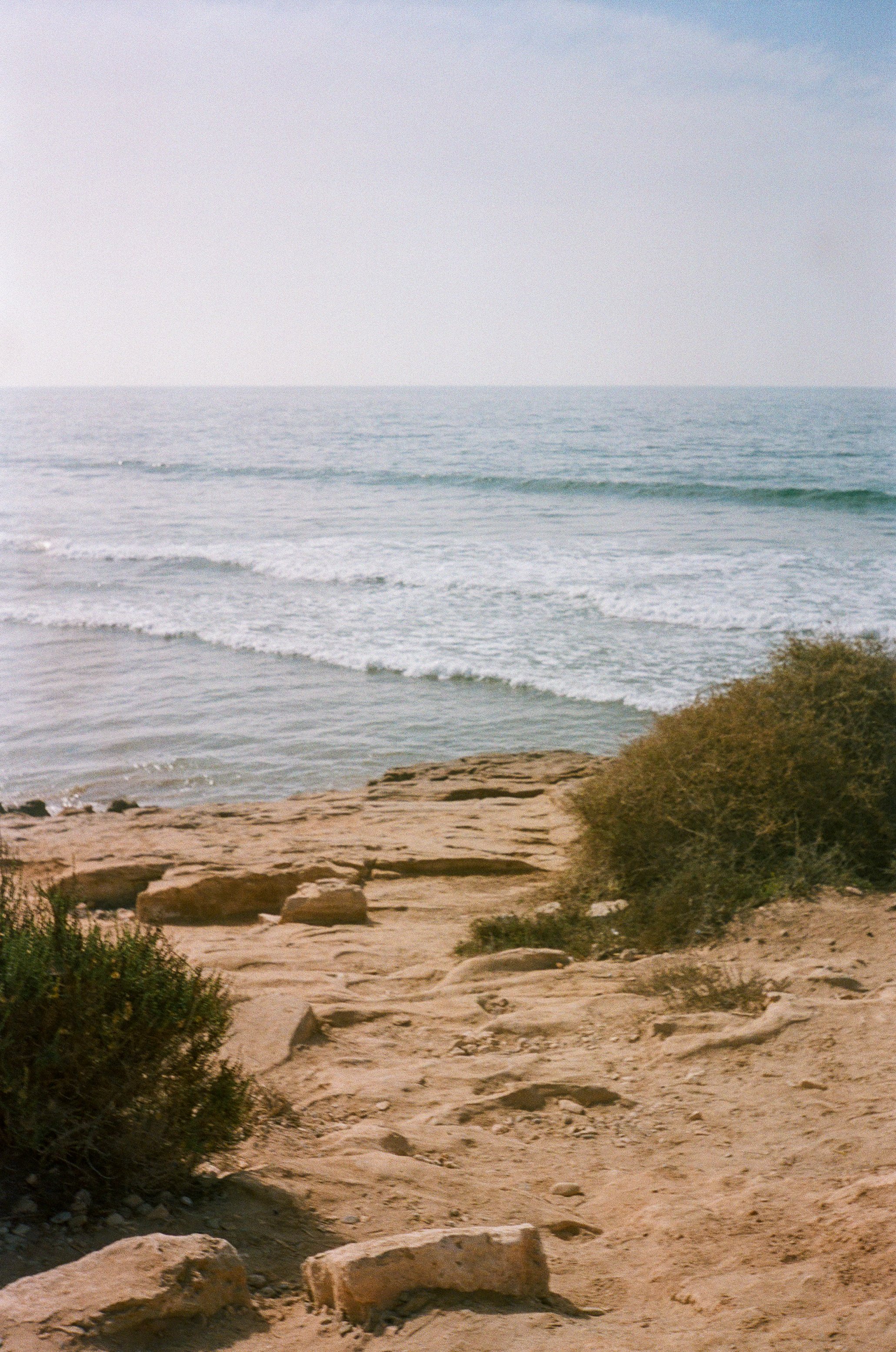A sandy pathway with rocks and vegetation leading towards the ocean, with calm waves and a clear sky in the background.