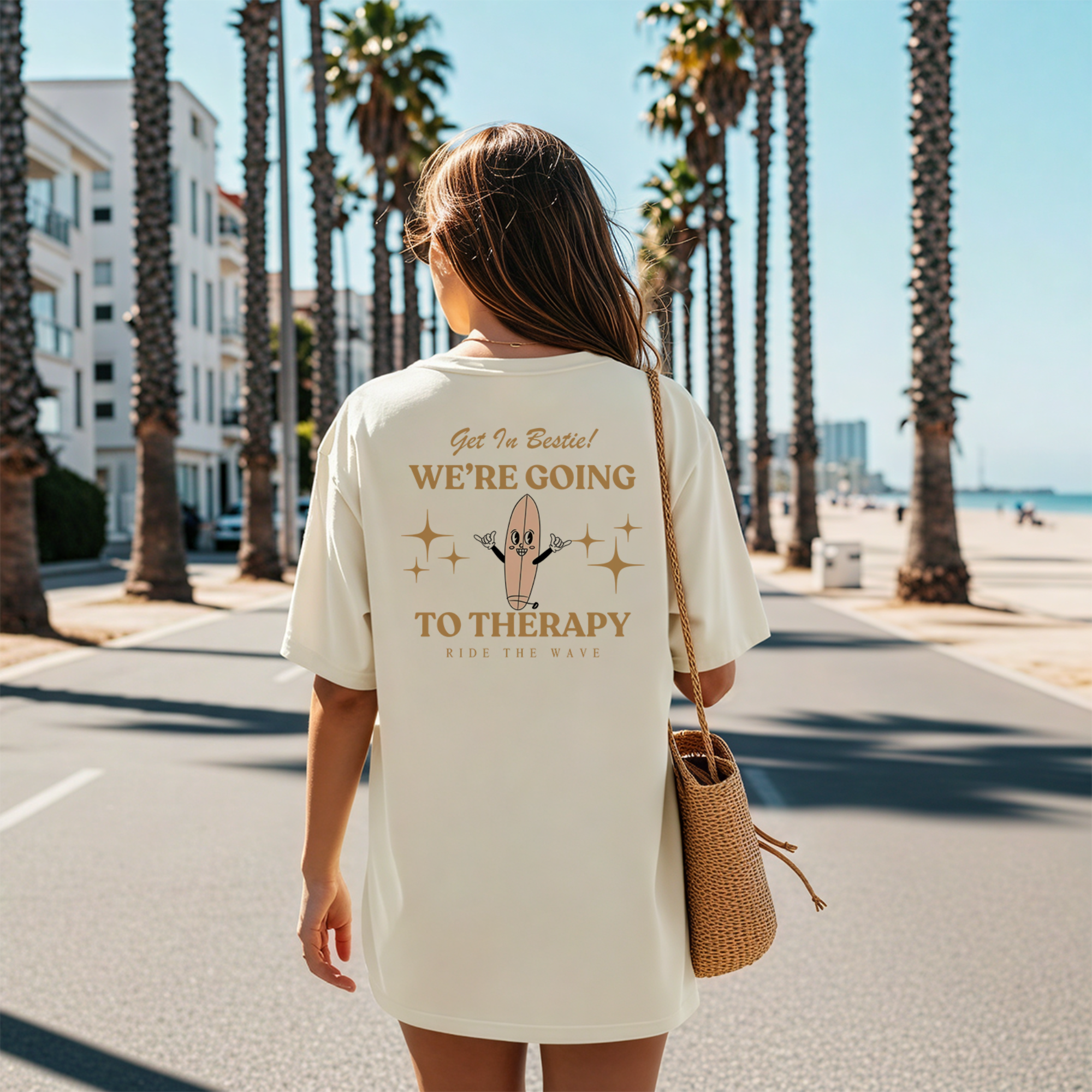 A woman walking on a sidewalk lined with tall palm trees and buildings in the background, wearing a cream-colored t-shirt with text and graphic, carrying a woven bag, during sunny weather.