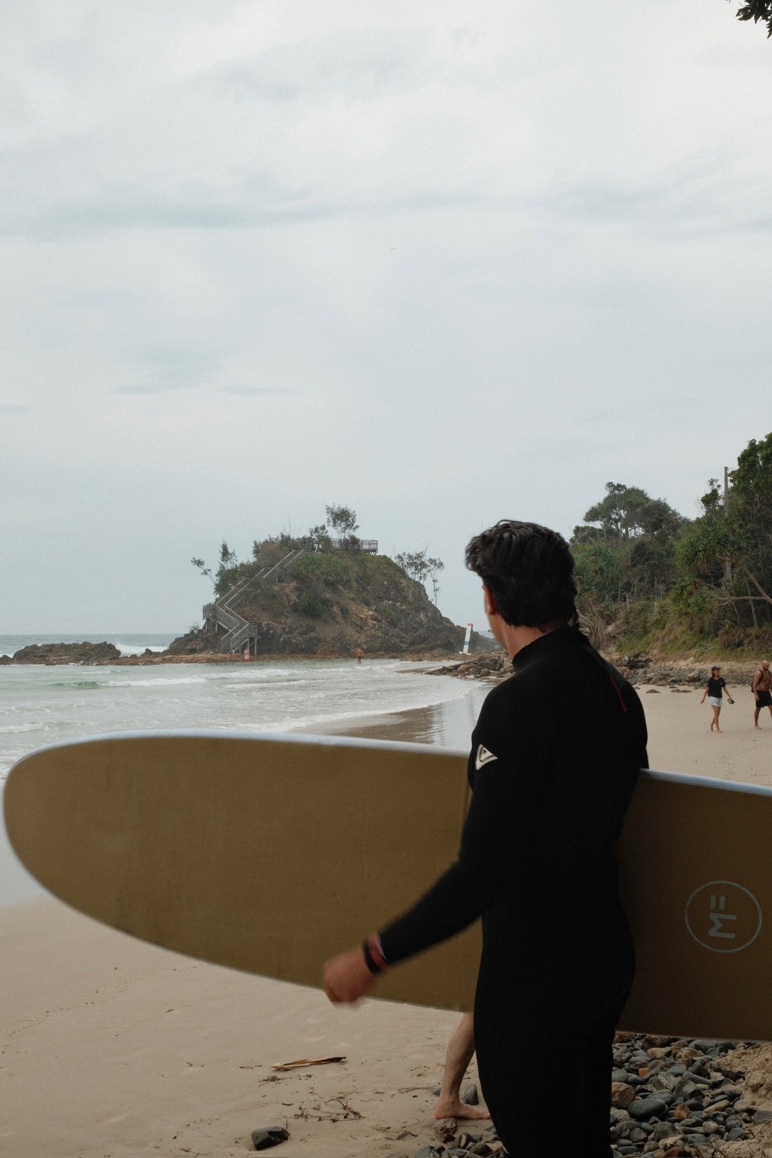 A person holding a yellow surfboard at a beach with a rocky hill in the background. The sky is overcast with some clouds, and other beachgoers are visible in the distance.