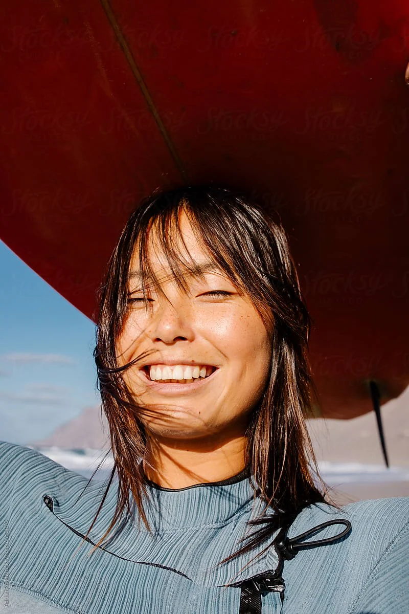 A woman smiling on a beach holding a surfboard over her shoulder.