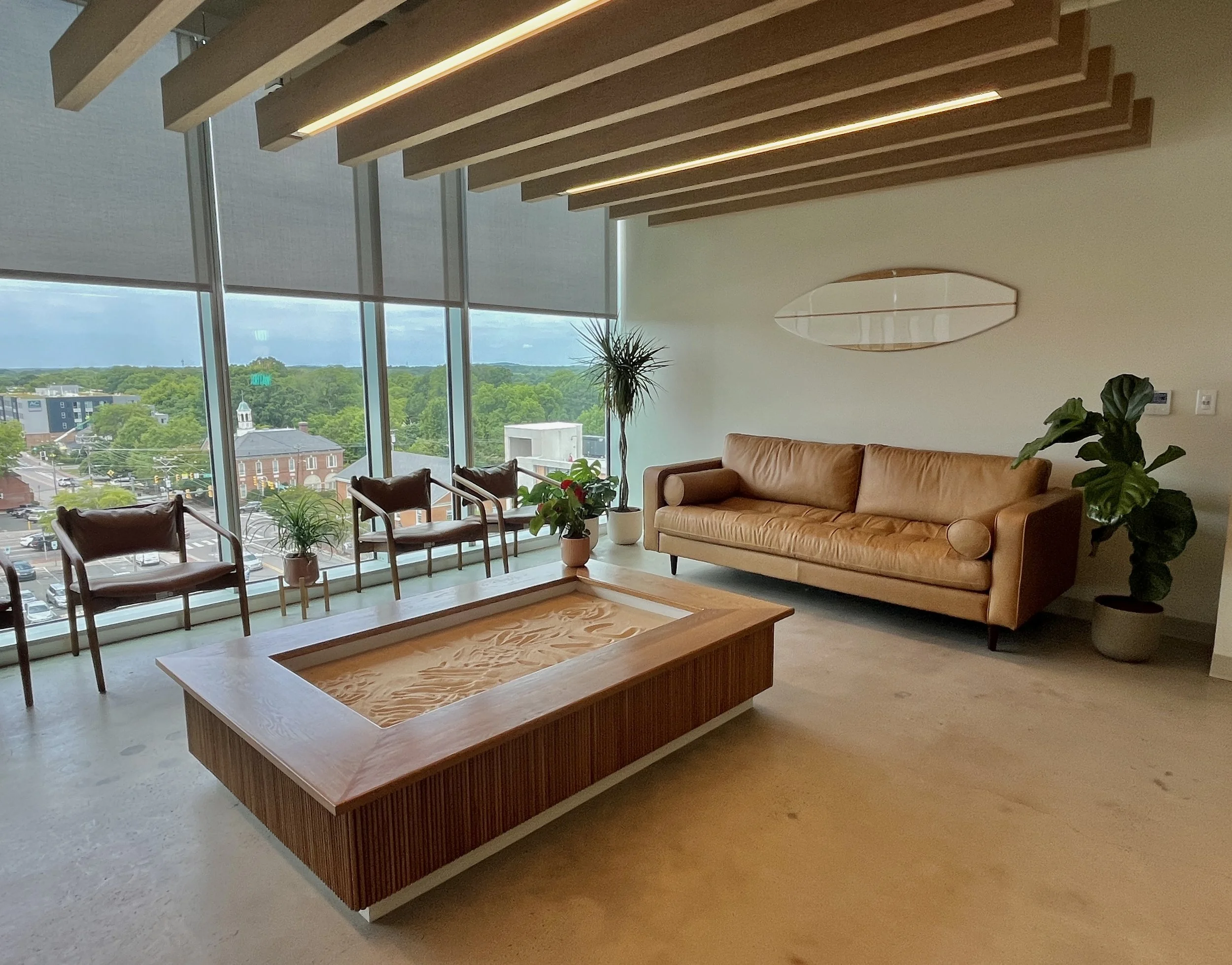 Modern office lounge with large windows, a leather sofa, wooden coffee table, and potted plants.