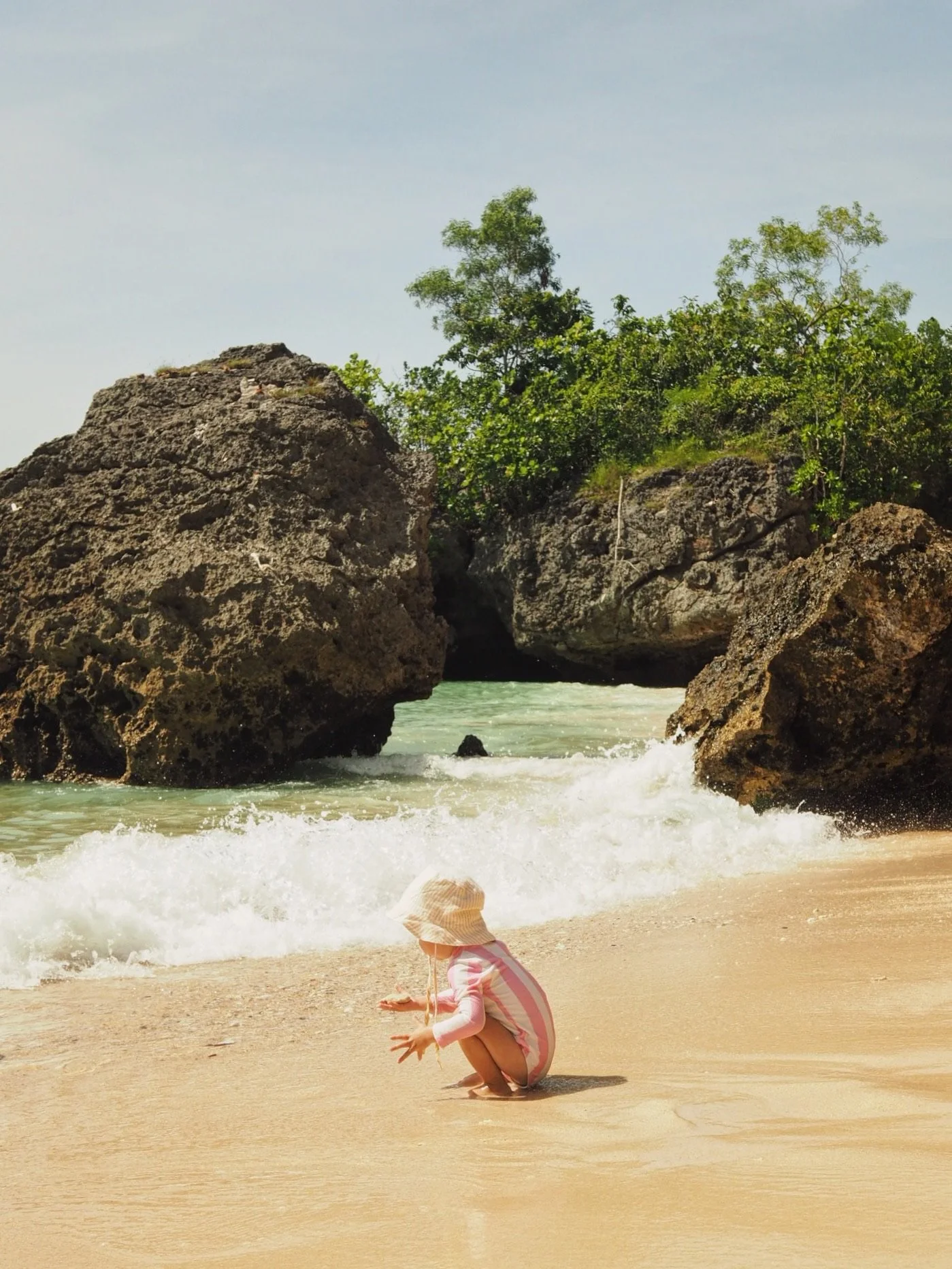 A child in a pink and white striped dress and a wide-brimmed hat kneels on the sandy beach near the water as waves break on the shore. Large rocks and green trees are visible in the background.