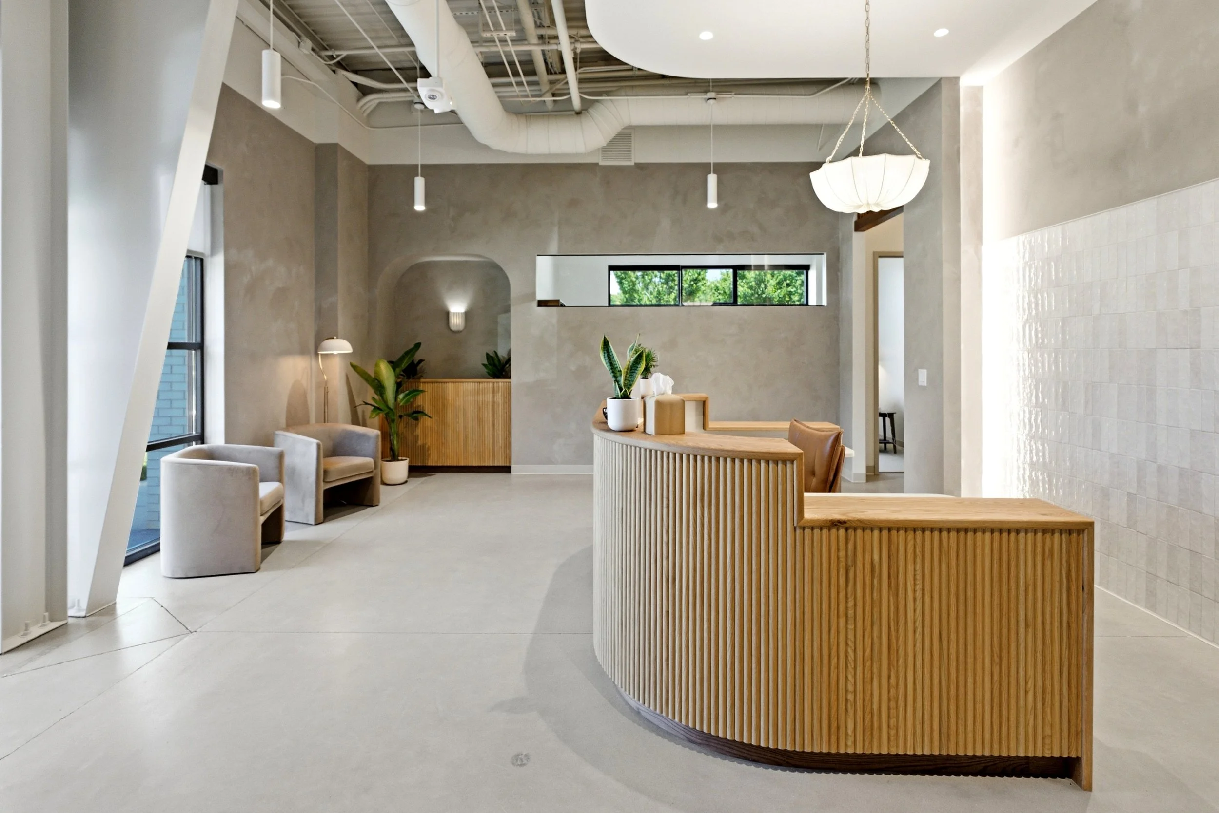 Modern interior space with light-colored walls, ceiling with exposed ductwork, hanging pendant lights, and a large window showing green trees outside. There is a curved wooden reception desk with potted plants, seating area with chairs, and abstract decor elements.