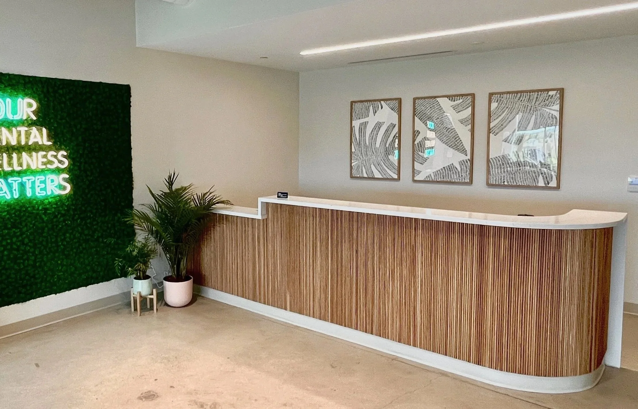 Reception desk with wood paneling, three framed palm leaf prints on the wall, potted plants, and a neon sign that reads 'YOUR MENTAL WELLNESS MATTERS' on a green leafy background.