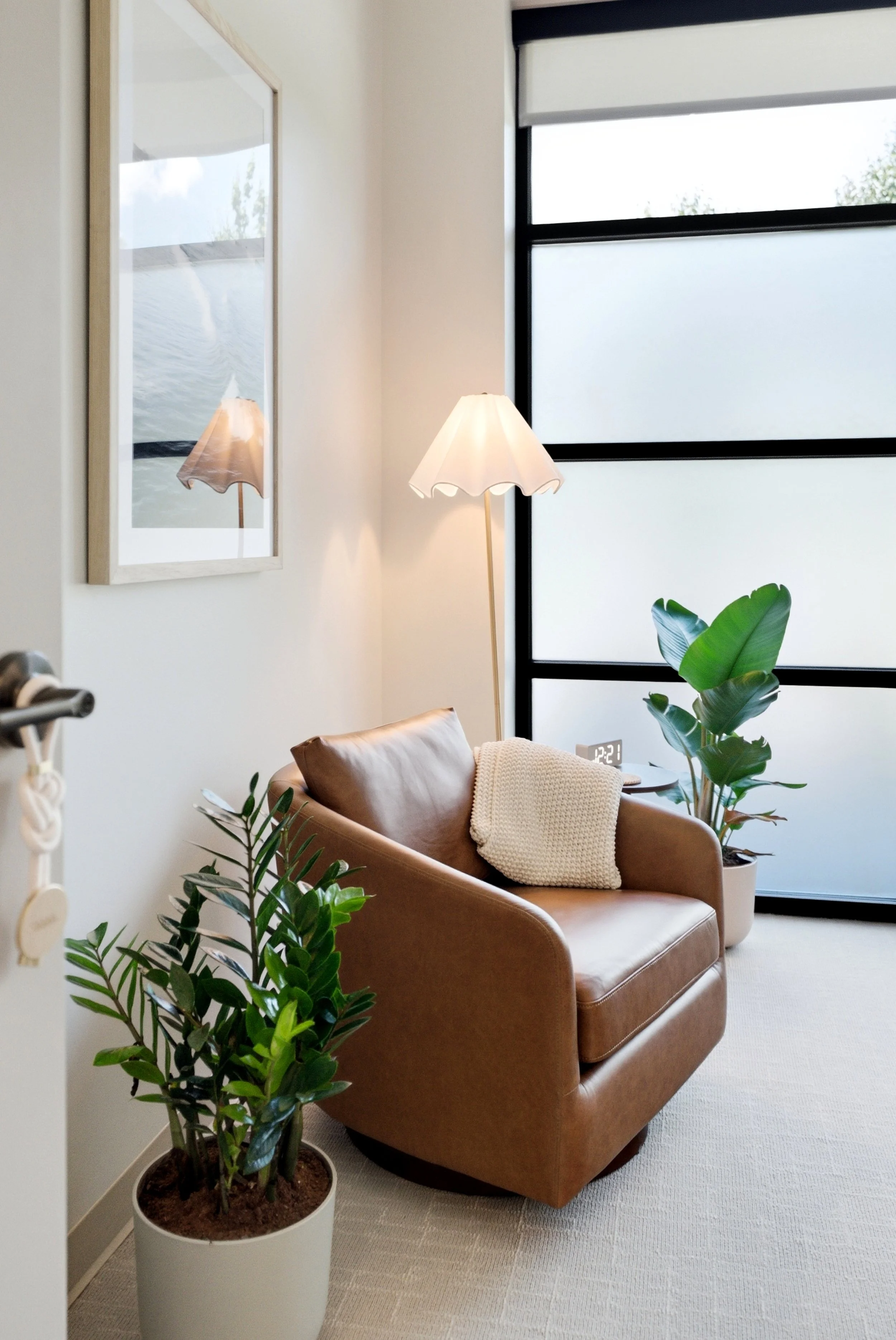 A cozy living room corner with a tan leather armchair, white blanket, and green plants, next to large frosted glass windows and a wall with framed artwork, illuminated by a floor lamp.