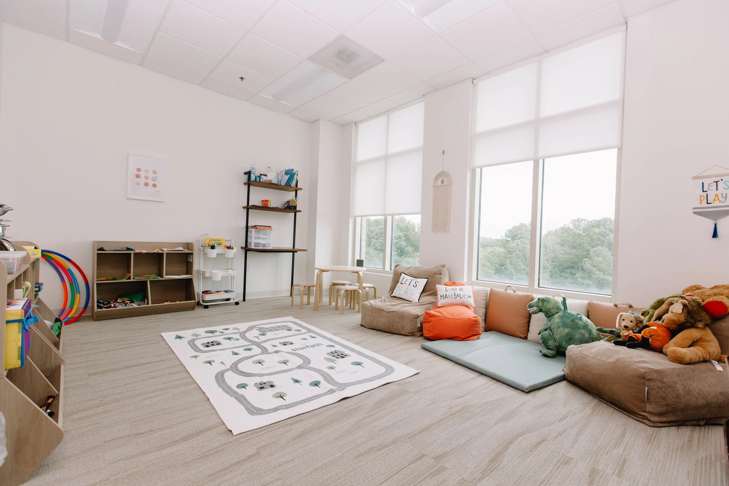 A spacious, well-lit indoor playroom with large windows, decorated with toys, plush animals, a play mat, and cozy seating.
