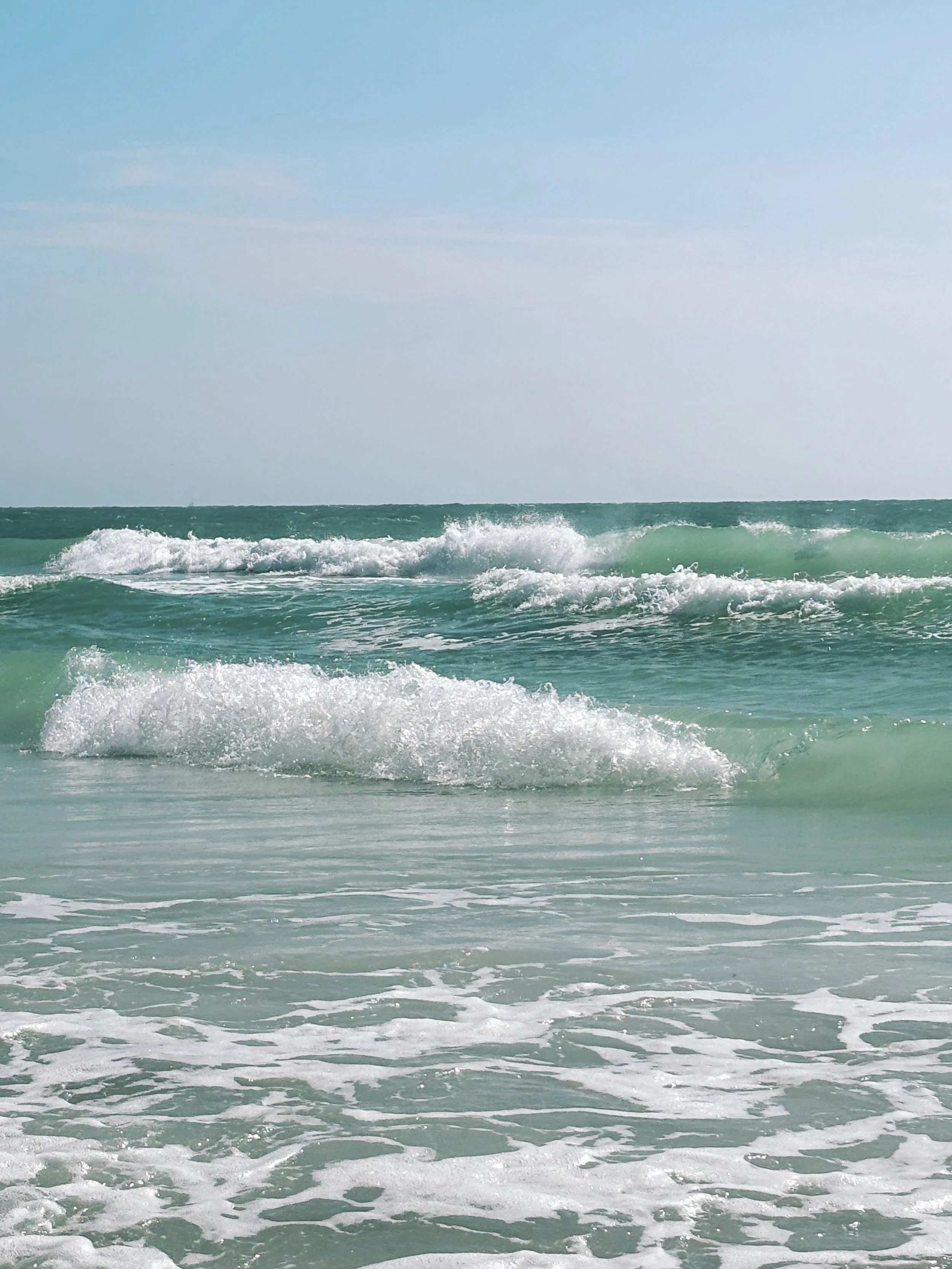 Ocean waves crashing on the shore under a light blue sky.