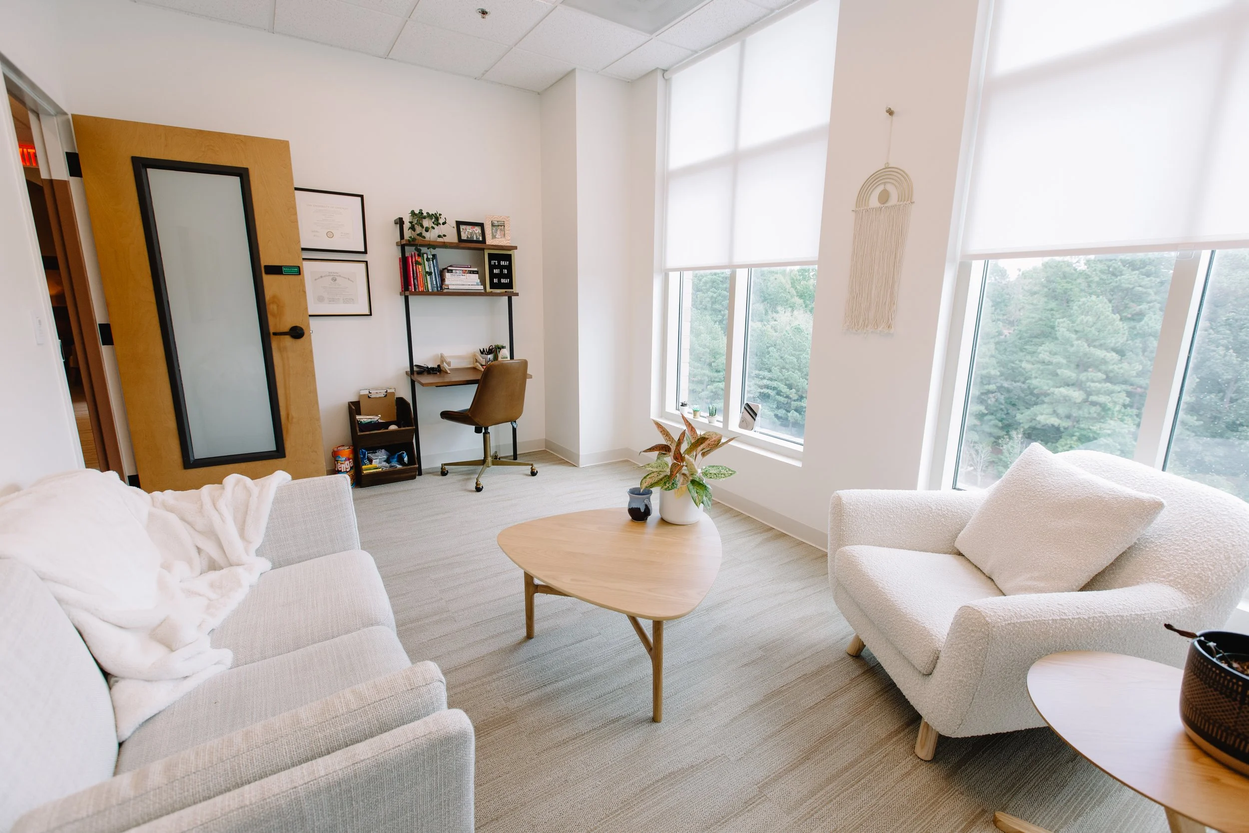 A bright, modern office with large windows, white walls, and cozy seating including a sofa and armchair, a small coffee table with potted plants, a wooden door, a bookshelf, and framed certificates on the wall.
