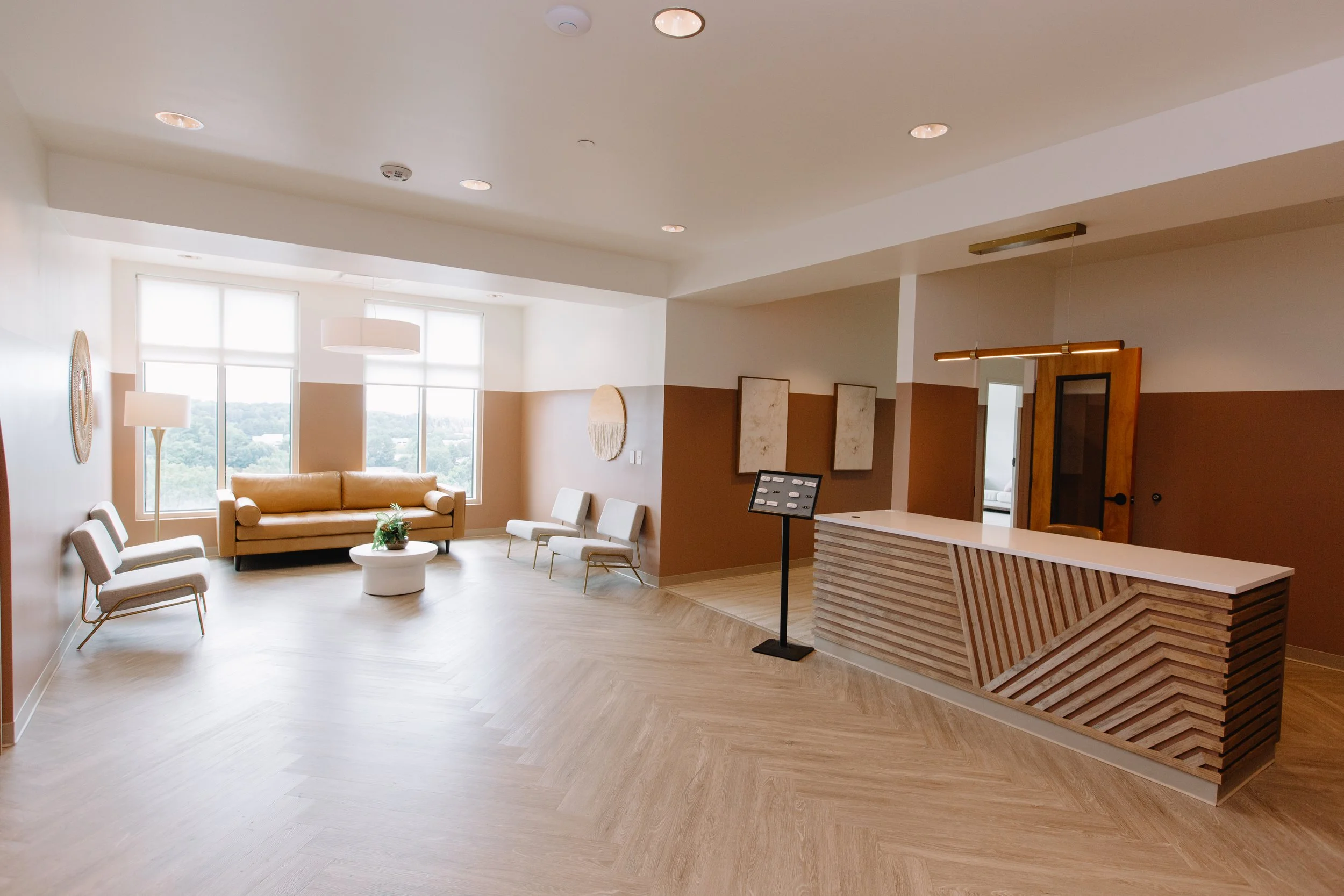 A modern, bright interior room with large windows, beige and white walls, a beige sofa, chairs, a white round coffee table with a plant, and a wooden reception desk.